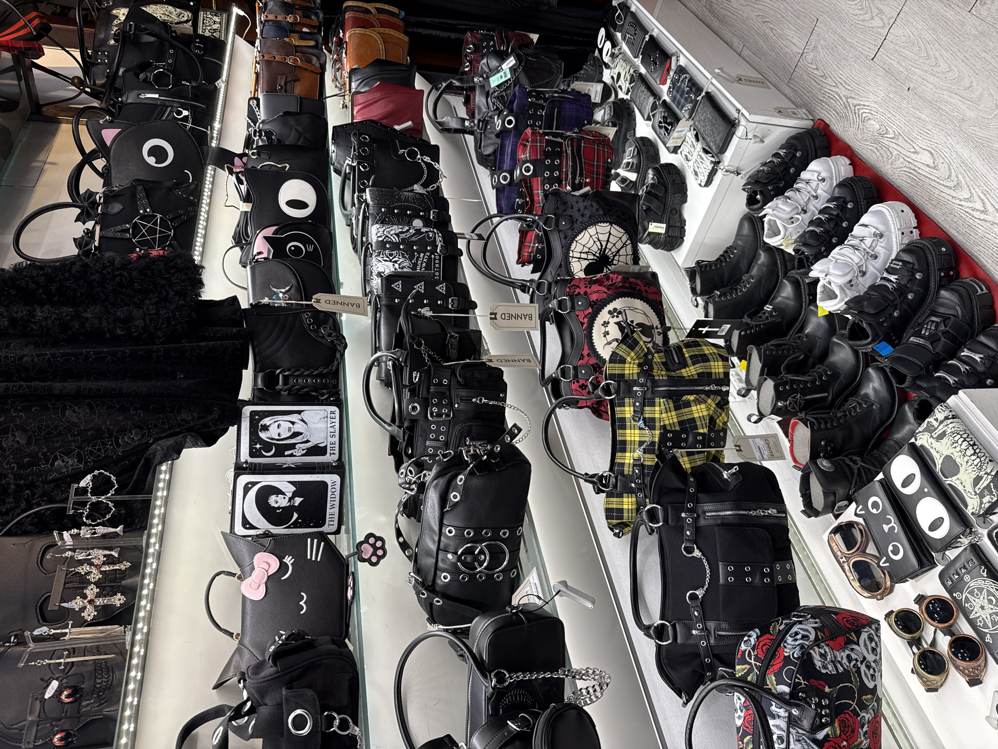 The image presents a well-organized display of alternative fashion items, primarily bags, wallets, and footwear, seemingly inside a retail store. The items are arranged neatly on shelves, with some bags bearing the brand name "Banned".
The bags exhibit a variety of styles, including those with gothic, punk, and kawaii influences. There are black bags adorned with studs, chains, and O-rings, as well as bags featuring character designs like cats with large eyes and Hello Kitty. Some wallets showcase tarot card-inspired designs and other gothic imagery.
Adjacent to the bags, a selection of footwear is displayed. The shoes and boots appear to be platform-style, with black and white color schemes and metallic accents. Other items visible include accessories like sunglasses, goggles, and jewelry. The environment suggests a store specializing in alternative fashion, possibly located in Madrid, Spain, given the implied context.