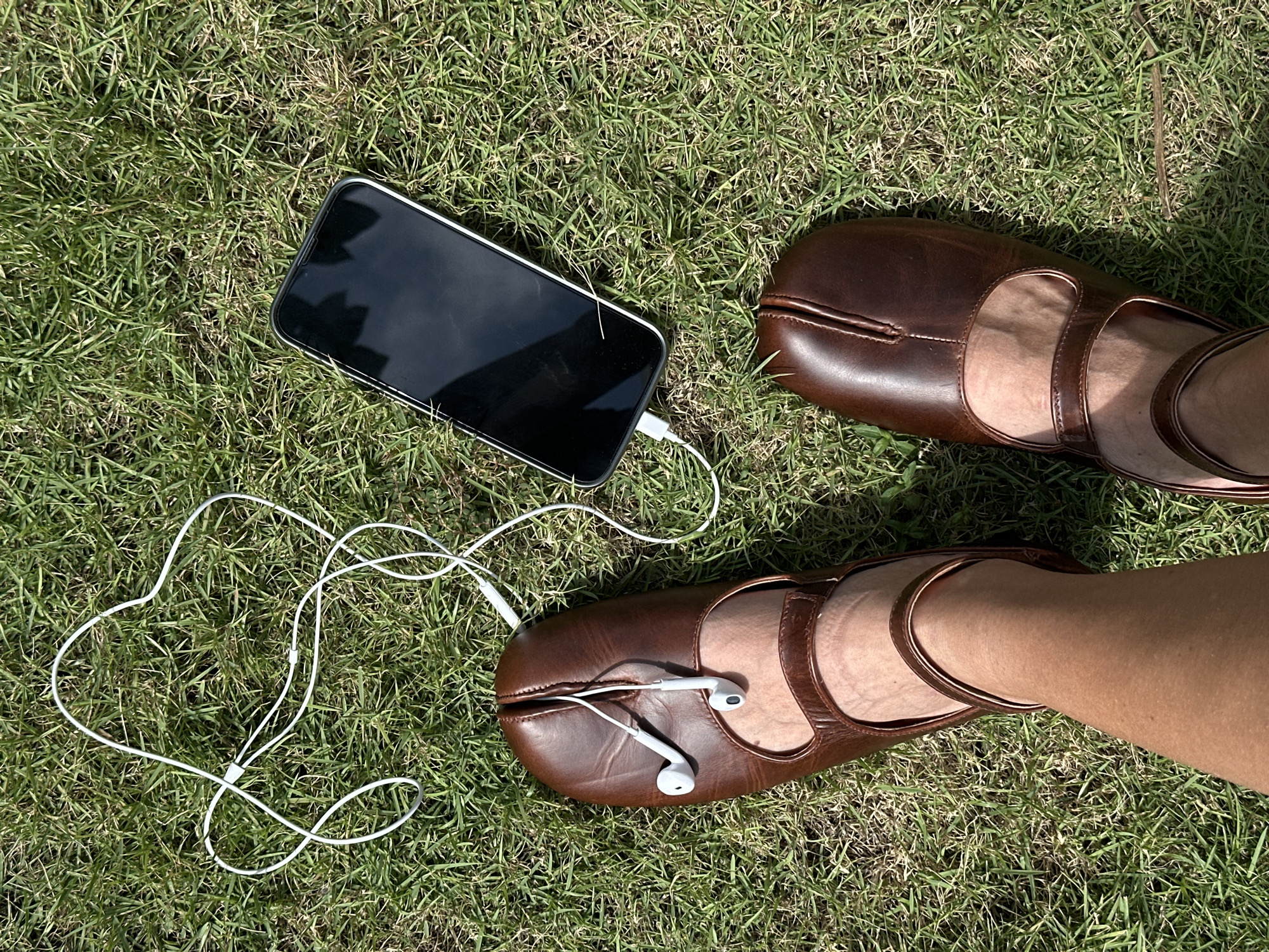 In North Kuta, Indonesia, on a sunny day, a person's feet wearing brown split-toe leather sandals are positioned on a lush green lawn. A smartphone is placed on the grass next to the sandals, connected by a white cord to earbuds. One earbud is dangling from the split of the left sandal, and the other is tucked into the sandal. The white cord of the earbuds forms a haphazard loop on the grass between the phone and the sandals. The grass appears dry in some patches, suggesting it may be late in the dry season.