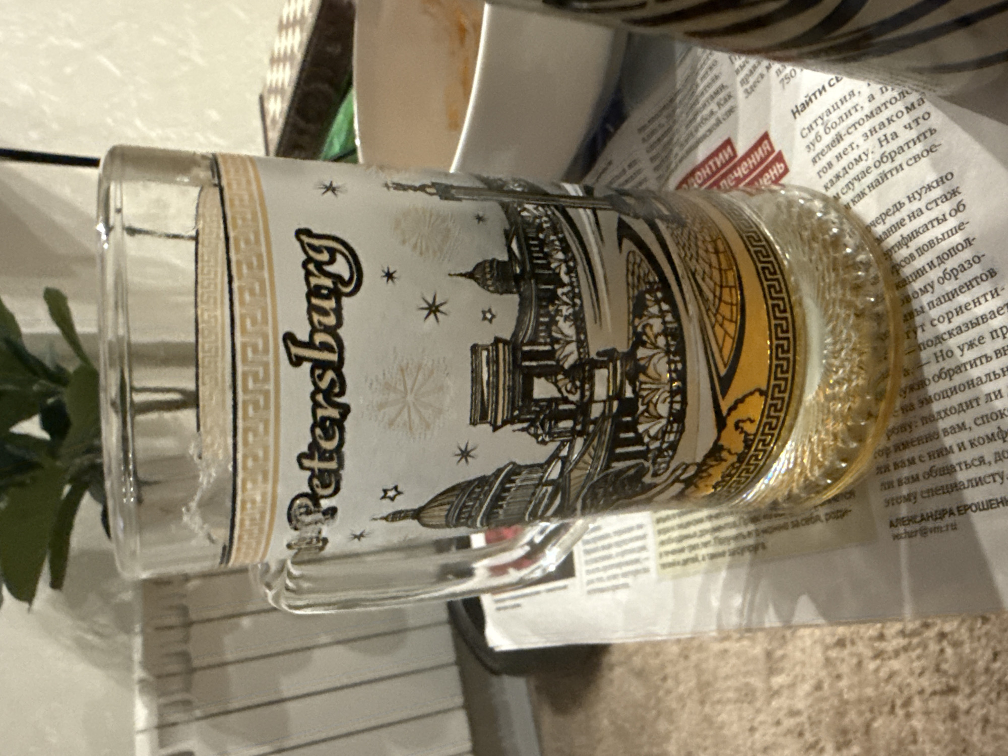 The image displays a clear glass mug, positioned horizontally on a light-colored surface. The mug contains a light yellow liquid, possibly a beverage. Its exterior features a decorative white design depicting an array of famous architectural landmarks characteristic of St. Petersburg, Russia, rendered in black and gold. The word "Petersburg" is prominently inscribed vertically in an ornate font on the left side of the design. The illustrations include recognizable structures such as the Kazan Cathedral, St. Isaac's Cathedral, and the Admiralty building, accented with small stars and snowflake-like motifs. A golden border with a Greek key (meander) pattern frames the top and bottom of the decorated band.
The mug is resting on an open newspaper, printed in Russian. Visible text on the newspaper includes sections related to "лечения" (treatment) and "одонтии" (odontology). Articles discuss topics such as finding a dentist when a "зуб болит" (tooth hurts) and advice on selecting specialists, emphasizing "стаж" (experience) and "сертификаты" (certificates). A name, "АЛЕКСАНДРА ЕРОШЕНЬ" (Alexandra Eroshen'), and an email address "cher@vme.ru" are also discernible. In the background, to the left, a green leafy plant is partially visible, and other household items like white containers and a dark, possibly patterned object, are seen. The setting appears to be a casual indoor environment.