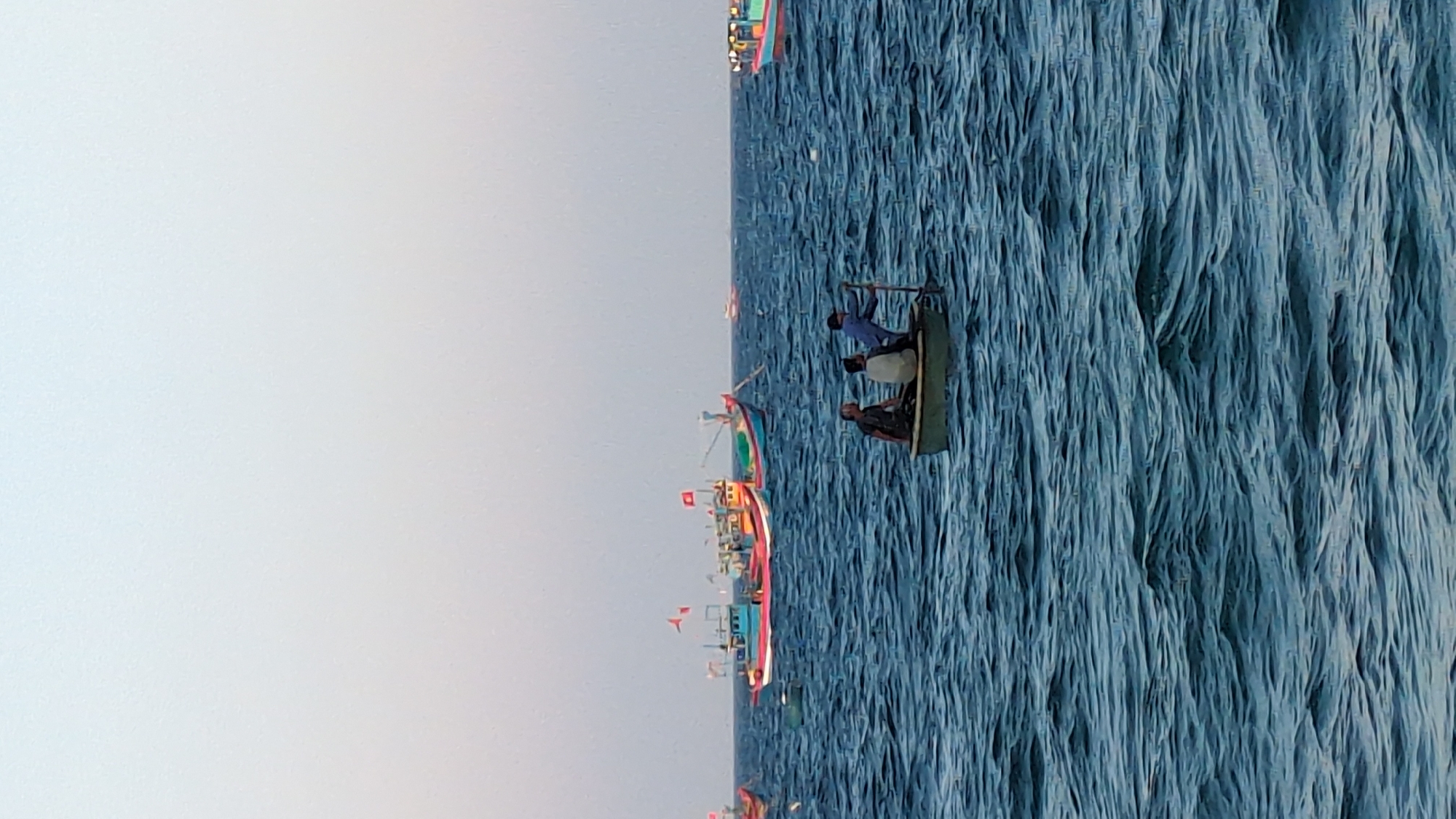 The image displays a marine scene in East Quy Nhon Ward, Vietnam, under a pale, hazy sky. The foreground is dominated by choppy, dark blue-green water. In the midground, a small, dark-colored, rounded traditional Vietnamese basket boat (thúng chai) contains three adult males. One male, dressed in a dark short-sleeved shirt, is seated on the left. Another male, wearing a light-colored long-sleeved shirt, is seated centrally. A third male, clad in a dark blue long-sleeved shirt, stands at the stern, operating a long wooden oar or pole, propelling or steering the vessel. In the background, several larger, brightly colored Vietnamese fishing boats are visible on the water. Two prominent vessels, one predominantly red and blue with a visible Vietnamese flag (red with a yellow star), and another primarily yellow, blue, and green, are positioned to the left. Additional smaller, less distinct vessels are scattered across the distant horizon.