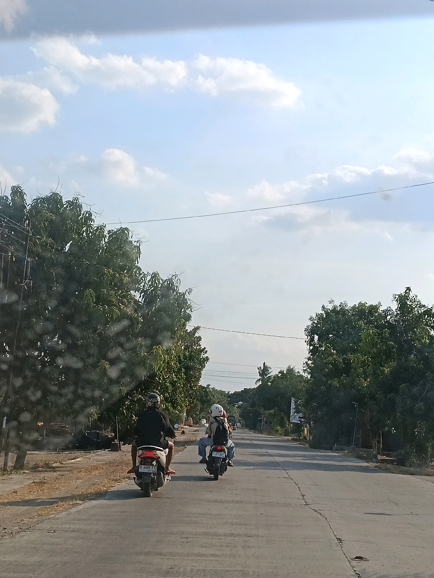 This is an outdoor, eye-level, medium shot of two people riding motorcycles on a paved road in Kutukan, Indonesia. The sky is bright blue with scattered white clouds, and the sun is casting shadows on the road. The road is lined with trees on both sides. The motorcycle in the foreground is black, and the rider is wearing a black jacket and a helmet. The motorcycle in the background is white, and the rider is wearing a white helmet and a backpack. The motorcycles are moving away from the viewer down the road. The overall impression is of a peaceful, rural scene.