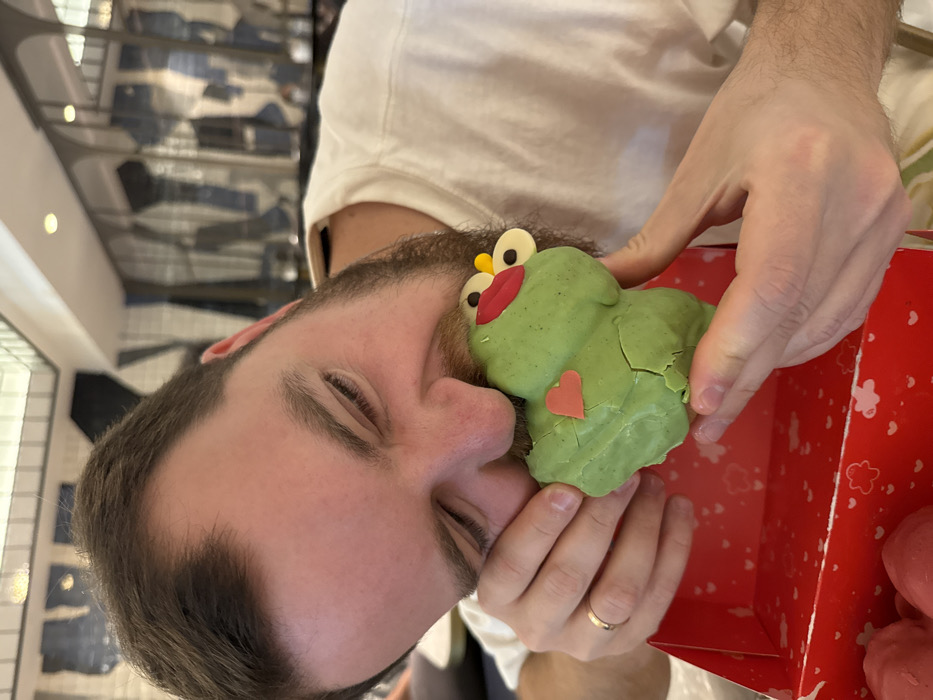 The image captures a close-up of an adult male in an indoor commercial establishment, specifically identified as a Demasié bakery/cafe in Barcelona, Spain. The subject, a man in his late 20s to early 30s with short brown hair and a beard, is actively biting into a large, bright green, frog-shaped pastry. His eyes are closed, and his mouth is wide open around the pastry, with a broad smile indicating enjoyment. He wears a plain white short-sleeved t-shirt and a thin gold band on the ring finger of his left hand.
The pastry is decorated with white chocolate eyes, yellow circular accents above them, red lips, and a small orange-red heart affixed to its chest. A portion of the pastry is missing from the bite, revealing a lighter interior, and its green shell shows several visible cracks. The pastry is held over a red cardboard box adorned with white and red heart patterns, implying a Valentine's Day or romantic theme. Other partially visible pink or red pastries are situated inside the box.
The background is blurred but shows a modern interior with a white tiled ceiling featuring integrated lighting. Behind the subject, a distinctive wall or partition, possibly viewed through a glass panel, displays abstract, geometric patterns in shades of blue and white, which resemble stylized representations of folded denim fabric. This specific decor is a characteristic design feature of Demasié bakeries.
