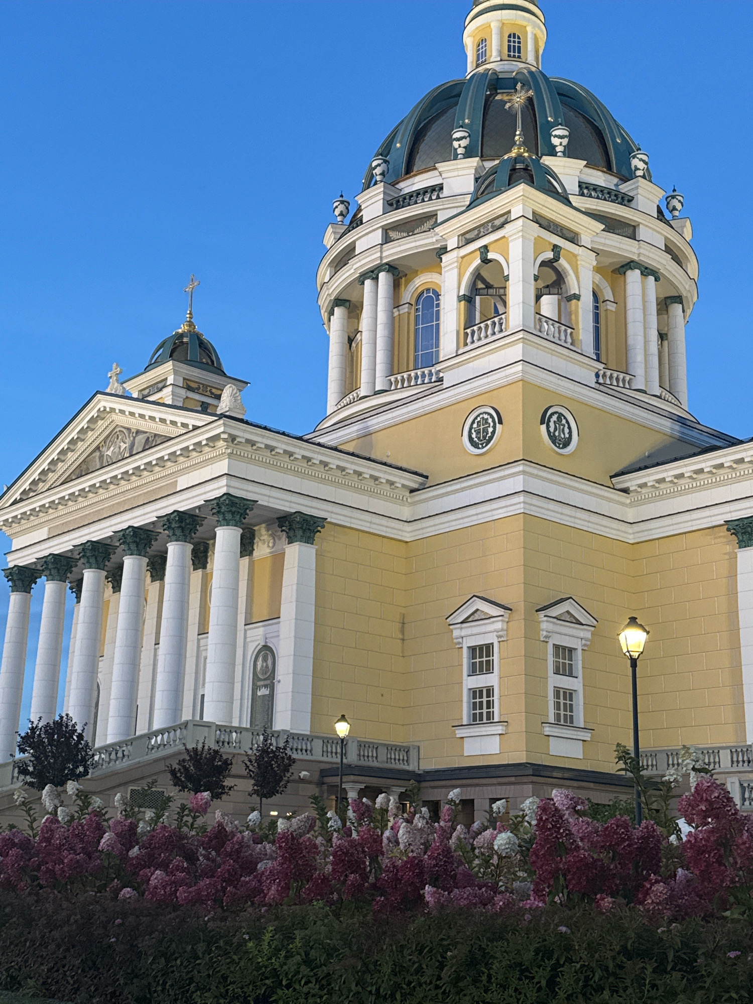 The image shows the Holy Trinity Cathedral in Chelyabinsk, Russia. It is a large, yellow building with white columns and a prominent dome. The cathedral is surrounded by a lush garden with pink and white flowers. The sky is clear and blue. The lighting suggests it is either early morning or late afternoon.