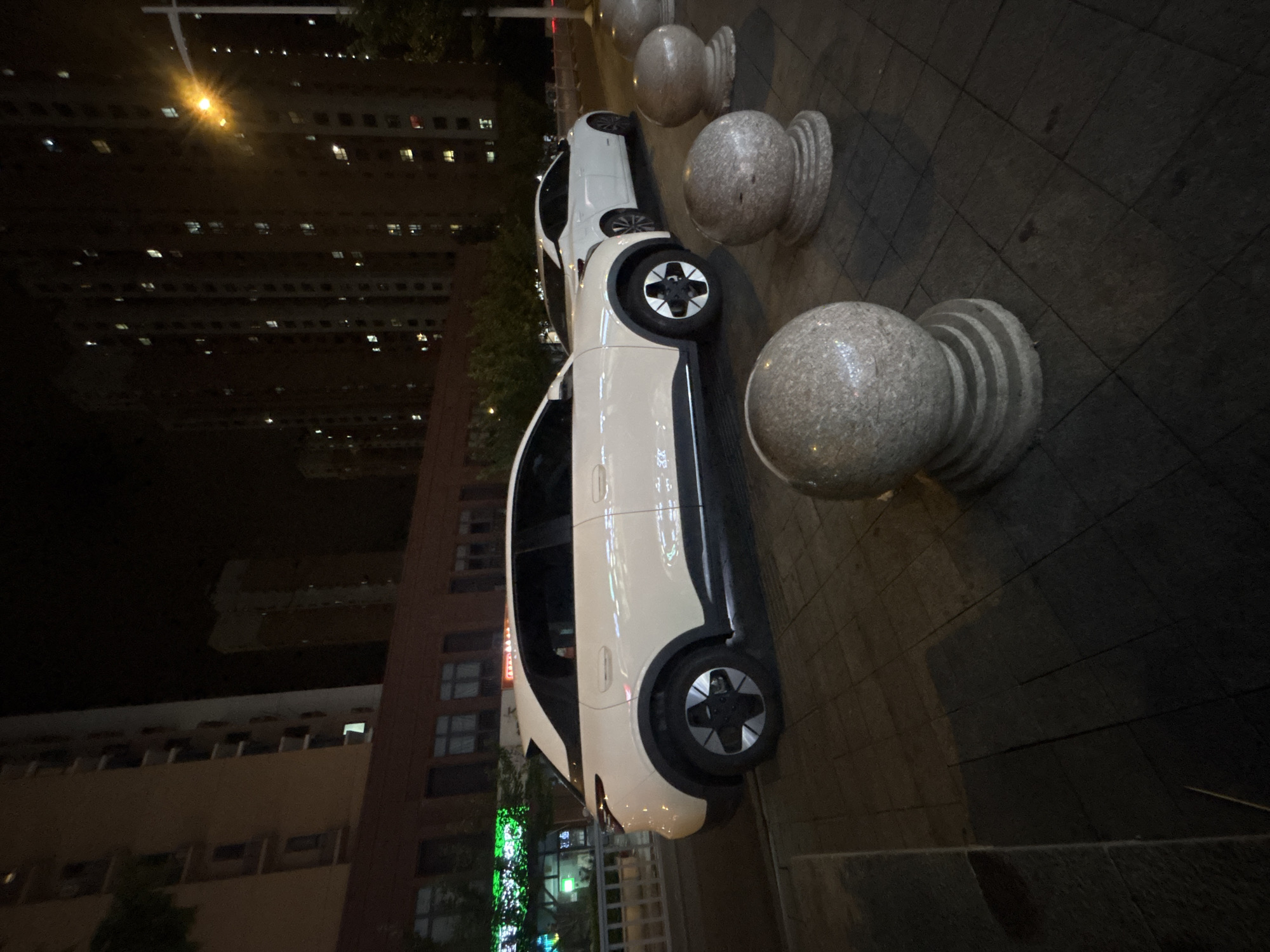 In the city of Suzhou Industrial Park, China, at night, two white cars are parked on a sidewalk alongside a building. The car in the foreground is a light-colored SUV with black trim around the wheel wells and a distinctive black wheel design. The second car, a white sedan, is parked further down the street behind the SUV. The sidewalk is paved with square tiles, and several round, stone bollards with tiered bases are placed along the edge of the sidewalk. In the background, tall apartment buildings with numerous lit windows are visible, indicating an urban residential area. A streetlamp with a bright, starburst-like glow is also present. The scene is illuminated by artificial lighting from the buildings and streetlamp.