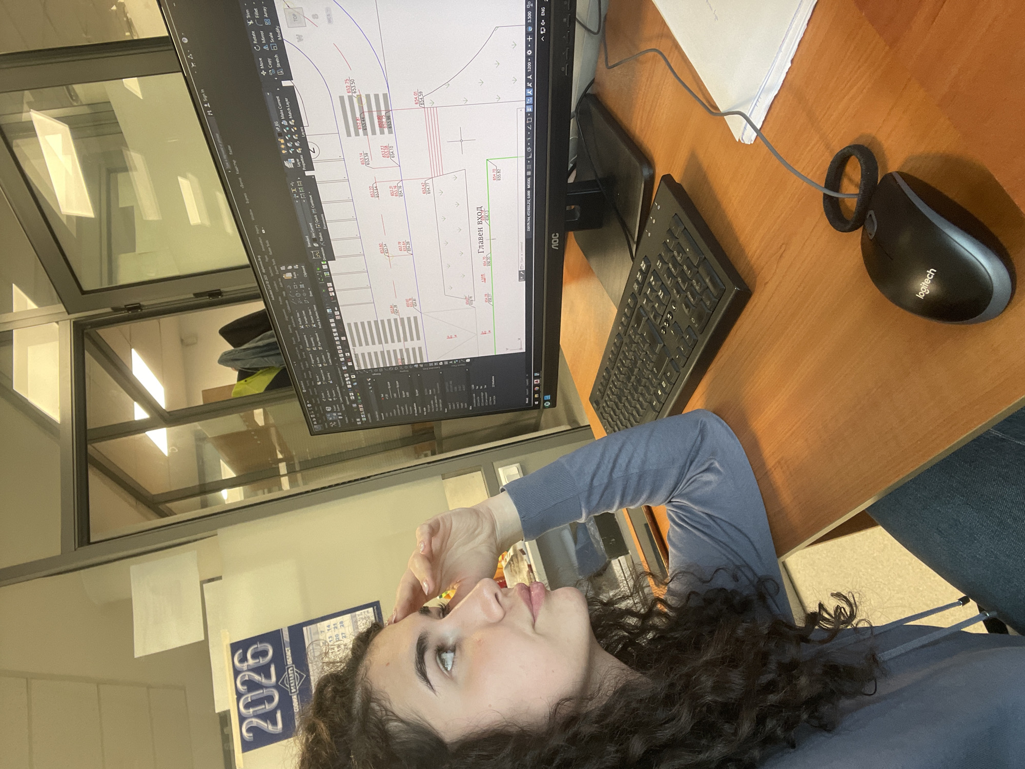 This media file captures a woman engaged in work within an office setting, likely located in Sofia, Bulgaria, as indicated by visible textual cues. The primary subject is a woman with prominent curly brown hair, dressed in a light blue long-sleeved top. She is positioned to the left of a wooden desk, looking upwards and to the right, with her right hand resting on her forehead, conveying a sense of deep thought or perhaps fatigue.
In front of her, a computer monitor, an AOC brand display, presents a detailed CAD (Computer-Aided Design) drawing. The screen showcases architectural or engineering plans, featuring numerical annotations and Cyrillic text including "Главен вход" (Main entrance). The software interface displays a range of CAD commands such as "Move," "Copy," "Rotate," "Trim," "Layers," along with coordinates and measurement data.
On the wooden desk to the right of the monitor, a black keyboard and a black Logitech wired mouse are arranged. A distinctive black ring is wrapped around the mouse's cable. The office environment is further defined by glass partitions in the background, which reflect overhead fluorescent lighting and elements of the room. A stack of white papers is also visible near the mouse.
A poster or calendar is affixed to a partition on the lower left, prominently displaying the year "2026" and Cyrillic text "СОФИЯ КИЛА ОБЛАСТ" (Sofia region), confirming the location context. The image depicts a focused, professional working atmosphere.