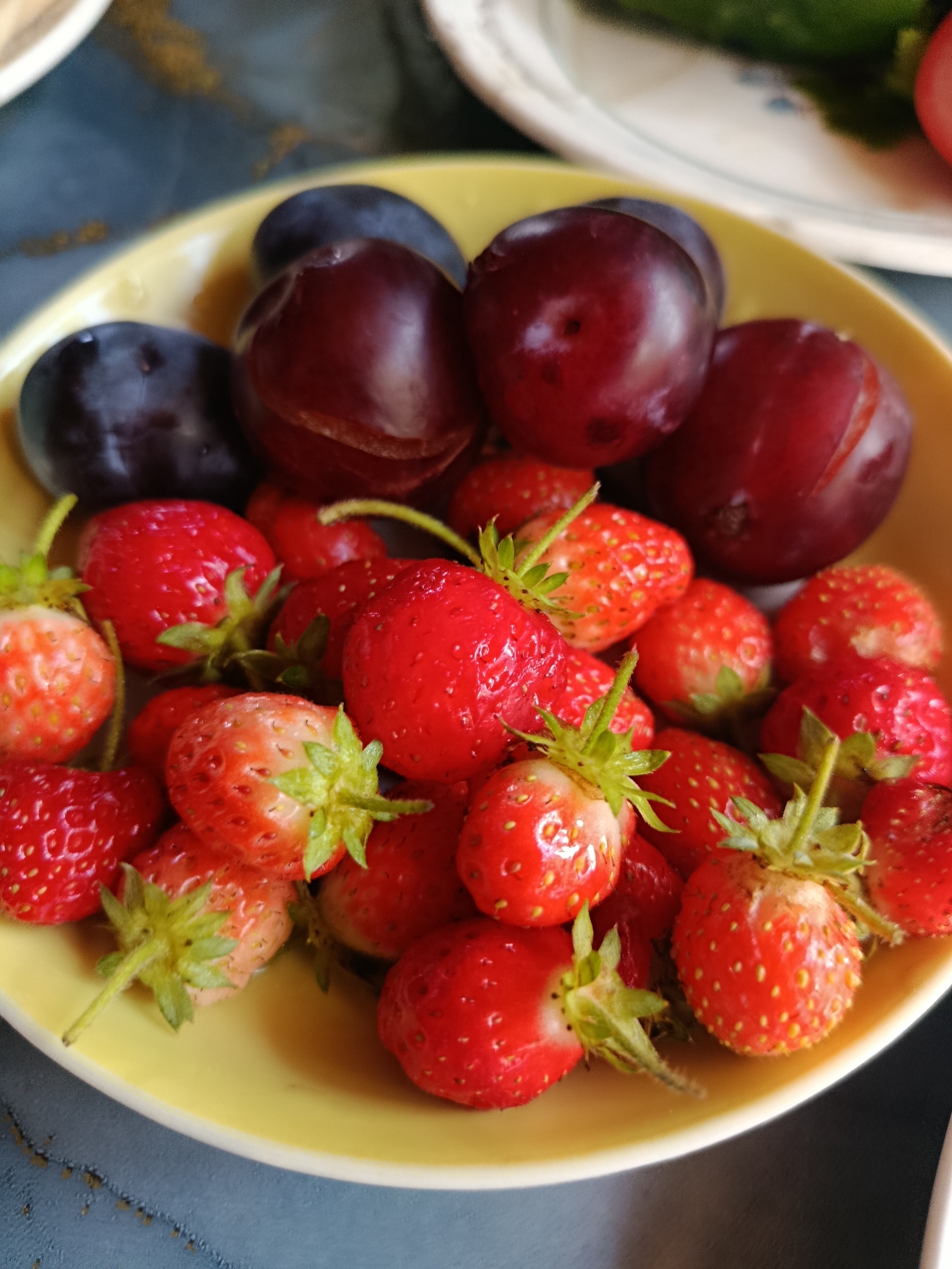 In a close-up shot, a yellow ceramic bowl contains an assortment of fresh fruits. The bowl is filled with plump, dark red plums and bright red strawberries. Some of the strawberries are whole, while others appear to be halved or partially submerged. The fruits are densely packed within the bowl, creating a visually appealing still life. The background of the image is blurred, but hints of other tableware and a dark surface suggest a domestic setting. The lighting appears to be natural, highlighting the vibrant colors and textures of the fruit. This image does not contain any specific geographical indicators that would place it in the city of Верея, Russia, or any other specific location.