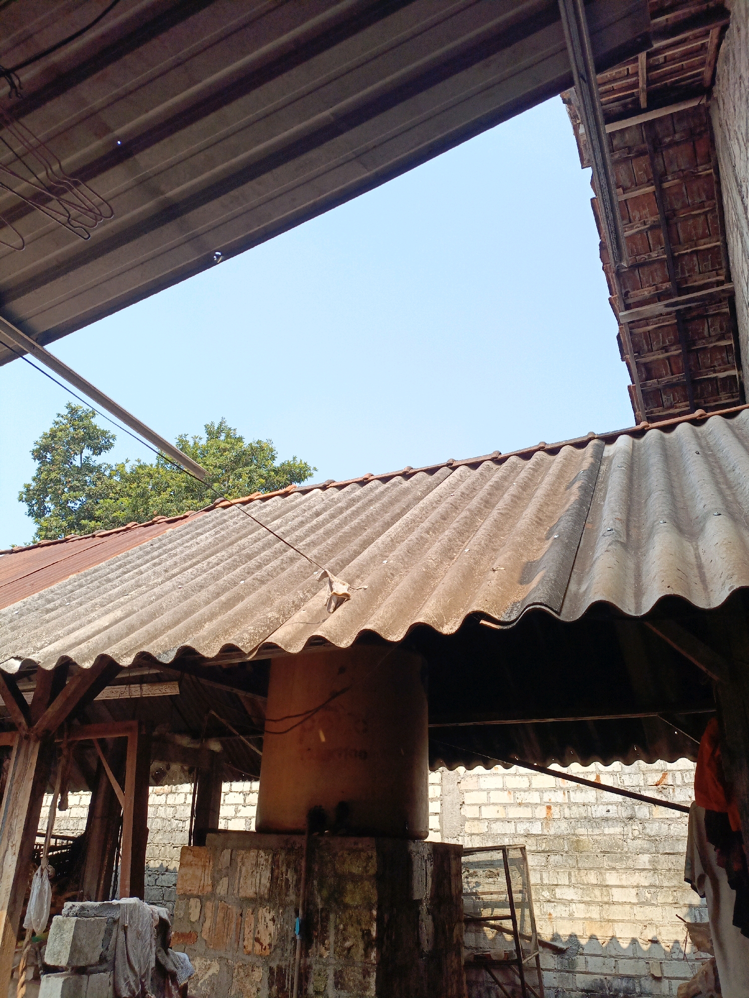 This is an outdoor, eye-level shot of a rustic structure with a corrugated metal roof in Jenu, Indonesia. The roof is weathered and shows signs of age. Beneath the roof, wooden beams are visible, supporting the structure. A large, cylindrical object, possibly a water tank, stands prominently in the center, made of an orange material. The background consists of a brick wall and some vegetation, including a large tree. The sky is clear and blue. The scene appears to be a functional area, perhaps for storage or a workshop, given the presence of building materials and the overall unrefined aesthetic. There are no people visible in the image.
