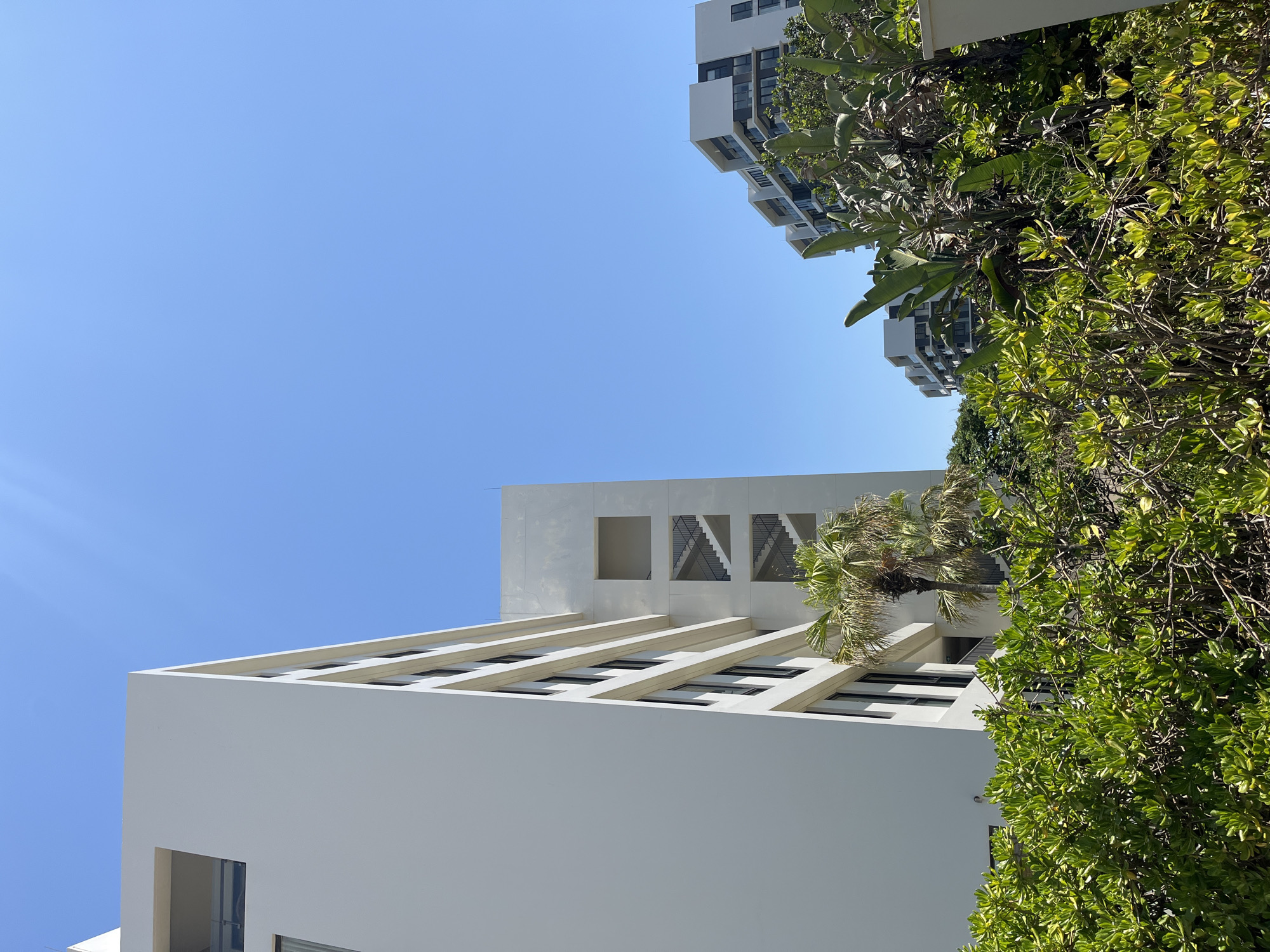 A low-angle shot shows portions of two modern buildings against a cloudless, bright blue sky. In the lower left, a white building with horizontal beams extending out from the main structure creates shadowed window spaces. A section above the beams features three square openings, two of which reveal the interior's staircase. To the right of this building, lush green foliage fills the right side of the frame. In the upper right corner, a taller building with multiple levels of balconies and windows is visible, though partially obscured by the foliage. The scene suggests an urban environment with tropical vegetation. The location is Đà Nẵng, Vietnam.