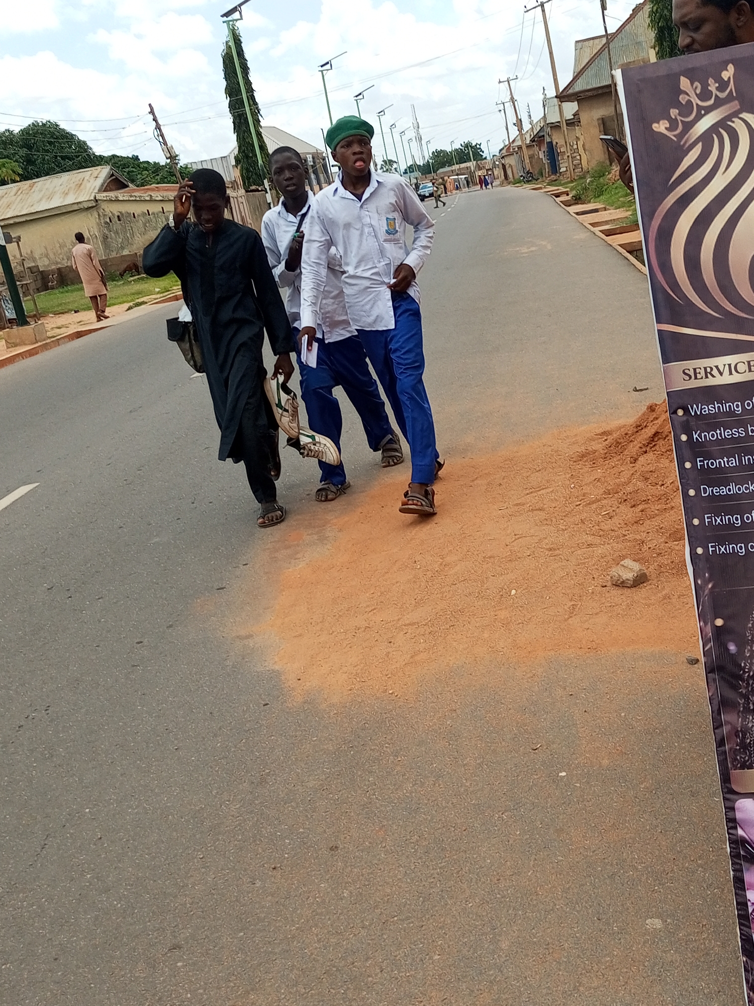 Three young men are walking on a paved road in Nassarawo, Nigeria. The man on the left is wearing a dark garment and carrying a white bag in his left hand and a pair of shoes in his right hand. The man in the middle is wearing a white shirt and blue pants, and the man on the right is wearing a white shirt and blue pants with a green cap. The man on the right is sticking his tongue out at the camera. The road is lined with buildings and trees. A banner with text and a logo is partially visible on the right side of the image. The sky is cloudy.