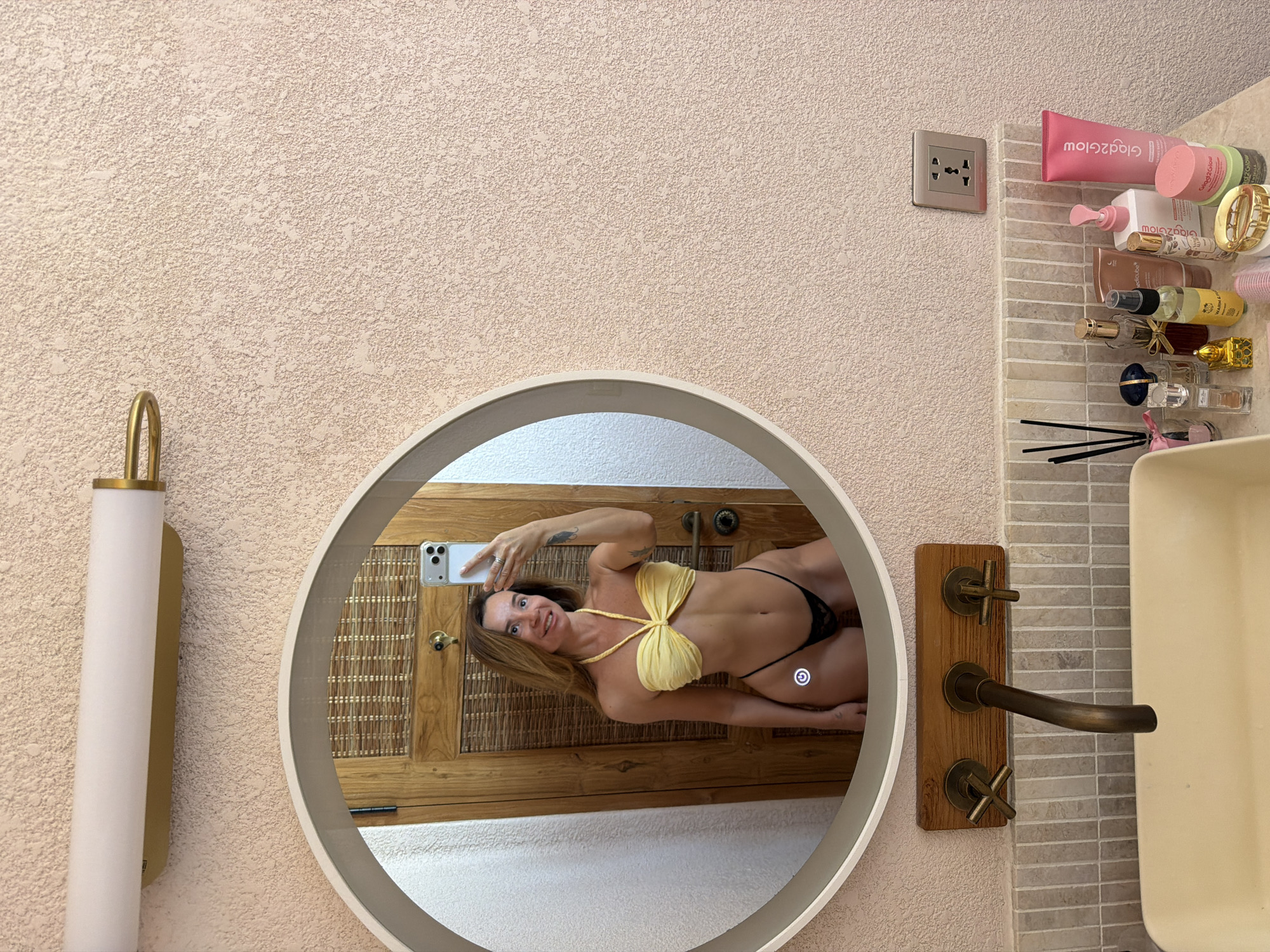 A woman is taking a selfie in a bathroom mirror. She is wearing a yellow bikini top and black bottoms. The bathroom has a textured pink wall, a round mirror with a light-up frame, a wooden door behind her, a sink with a bronze faucet, and various toiletries on the counter. The scene appears to be in Mengwi, Indonesia.