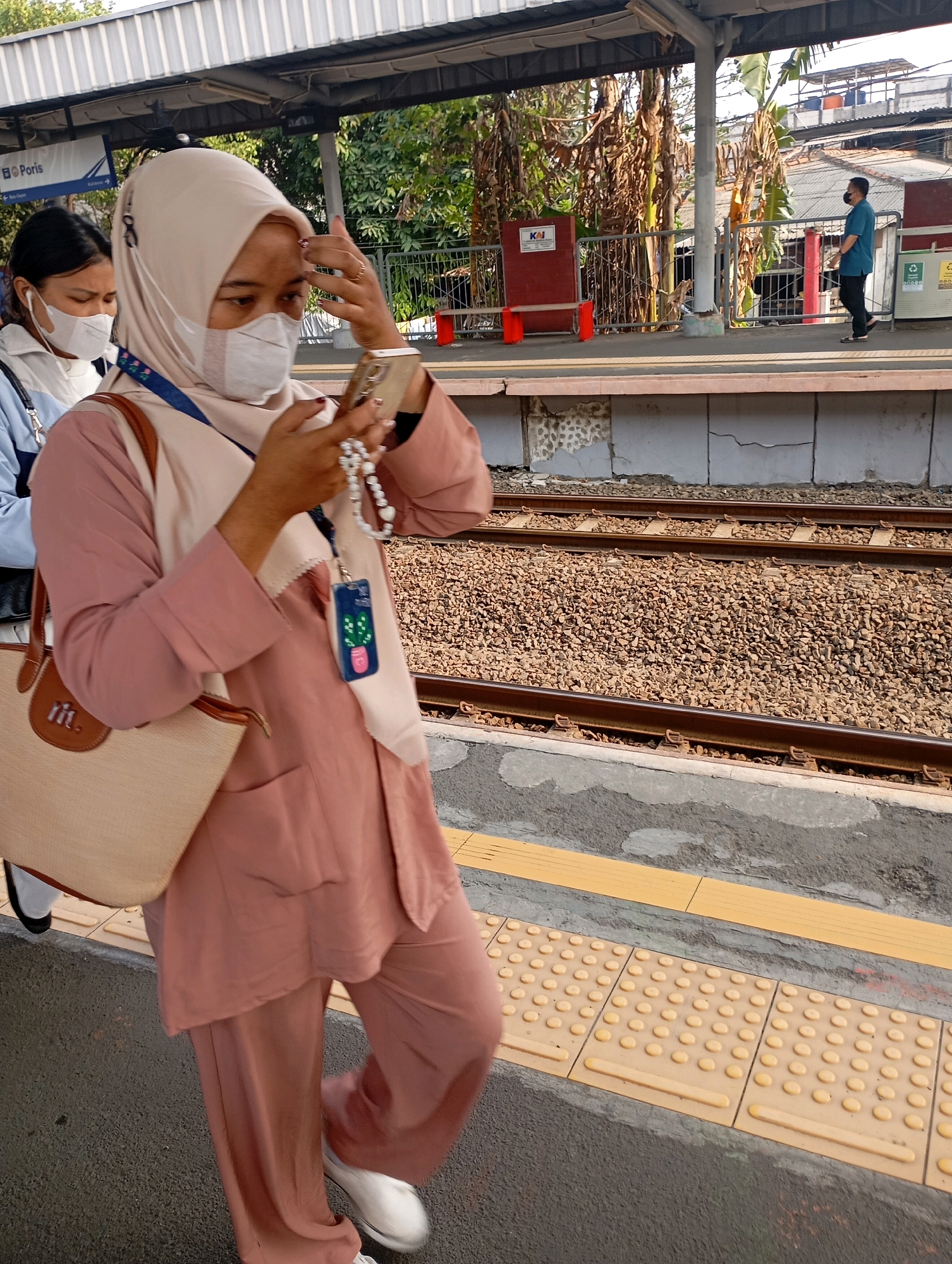 A woman wearing a light pink hijab and a light pink mask, along with a light pink long-sleeved shirt and matching loose pants, is standing on a train platform in Tangerang, Indonesia. She is holding a smartphone and a string of white prayer beads in her hands, appearing to be looking at the phone. A light beige tote bag with brown trim is slung over her left arm, and she is wearing white slip-on shoes. To her left, another woman wearing a white mask and dark clothing is visible. In the background, there are train tracks, a covered platform, and buildings. The platform has tactile paving. A man in a blue shirt and black pants is walking in the distance on the platform. The scene appears to be at a train station during the daytime.