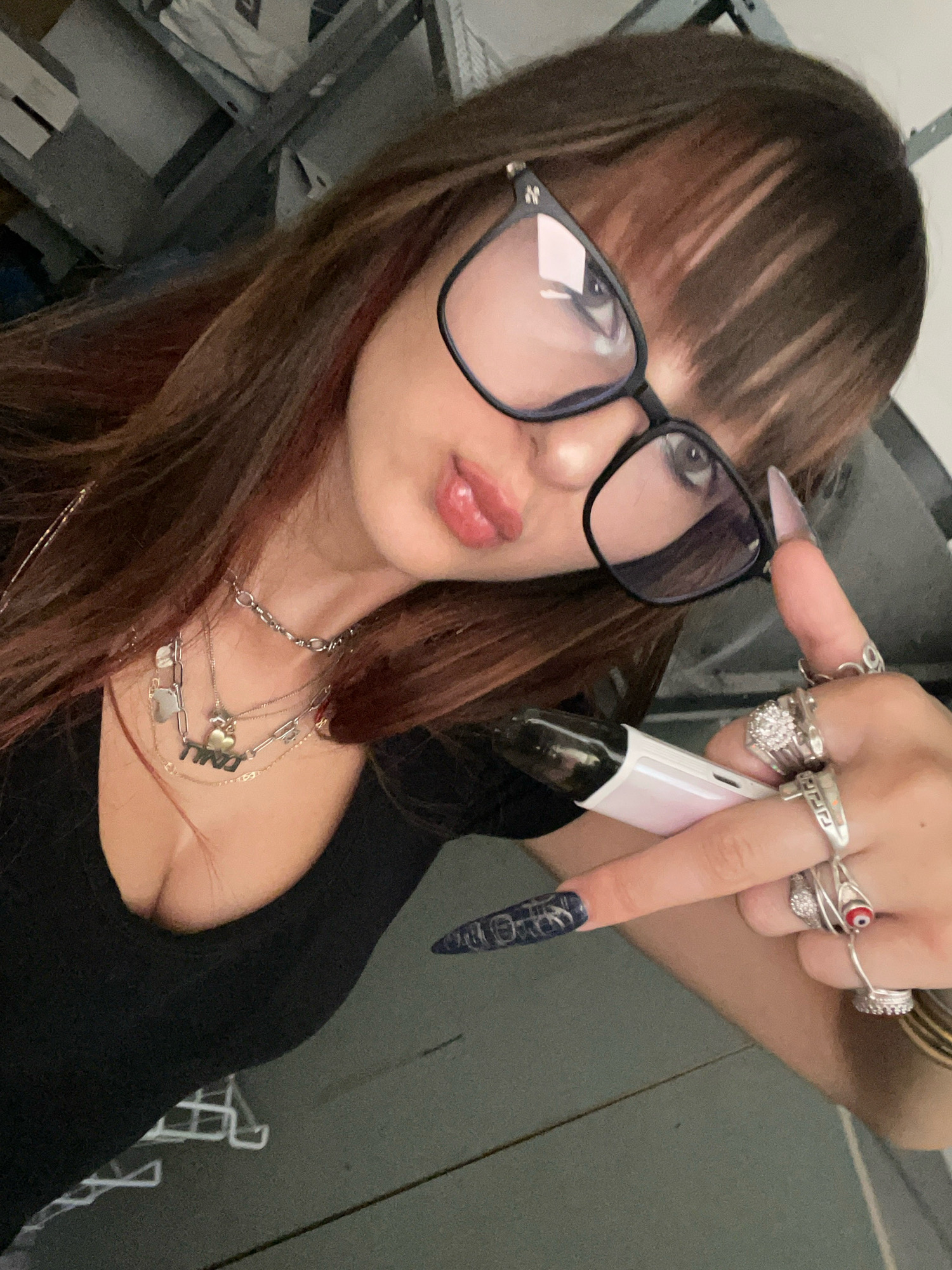 A young woman with long brown hair and bangs is taking a selfie. She is wearing large black-rimmed glasses and a black top. She has several necklaces on, including one with charms and another with the word "NINO" in black lettering. Her fingernails are long and painted with a dark blue design. She is wearing multiple rings on her fingers, including a large diamond-like ring and a ring with a red evil eye. She is holding a white and pink vape pen in her right hand and is making a peace sign with her left hand. The background appears to be an indoor setting with shelves and some bags, suggesting it could be a storage area or a messy room. There is no indication of the specific location within Raanana, Israel, but the overall environment suggests a casual, personal space.