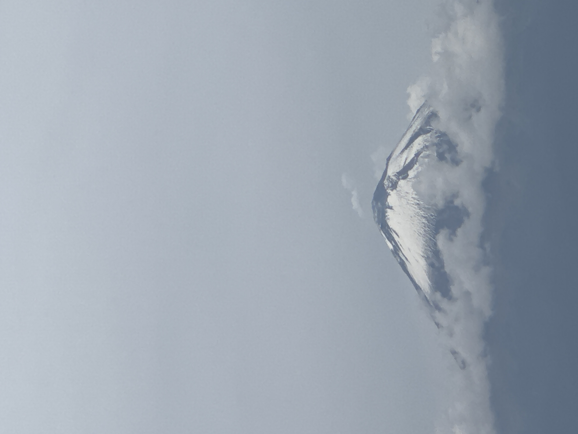 An overcast sky dominates the upper portion of the image, with a large, snow-capped volcano emerging from a bed of clouds in the lower half. The volcano's peak is conical and appears to be actively smoking, with a faint plume rising from its summit. The snow cover is patchy, revealing dark rock beneath. The clouds partially obscure the lower slopes of the volcano, creating a sense of depth and mystery. The overall impression is one of natural grandeur and a hint of volcanic activity. The location is San Bernardino Tlaxcalancingo, Mexico, a region known for its proximity to several volcanoes.
