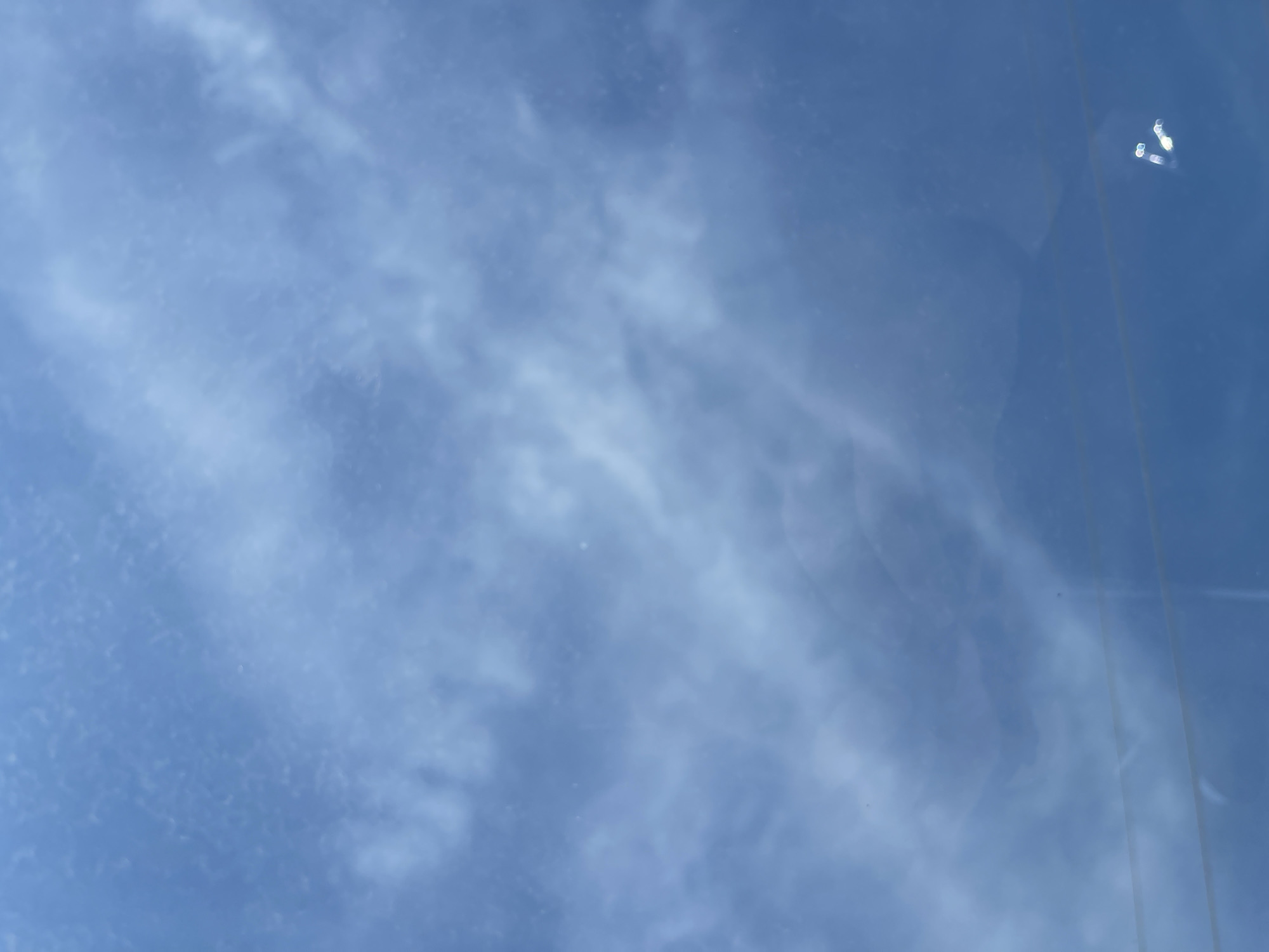 The image shows a blue sky with streaky cirrus clouds. The image is taken through a window, as evidenced by reflections and a linear distortion along the right edge. The location in the city of Kueda, Russia, is unknown and cannot be determined from the visual information.