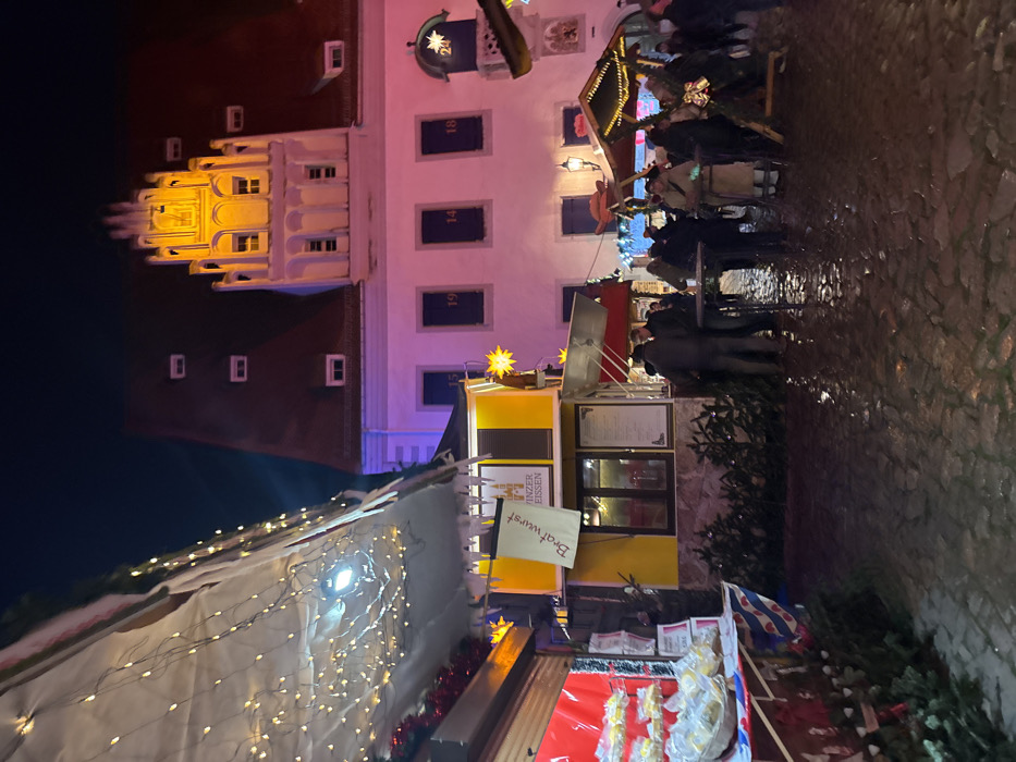 A nighttime outdoor shot shows a Christmas market scene. A building stands tall with brightly lit windows. Market stalls are decorated with lights and offer food like bratwurst. People are milling about. The ground is wet, reflecting light. The photo is taken from a slightly low angle.