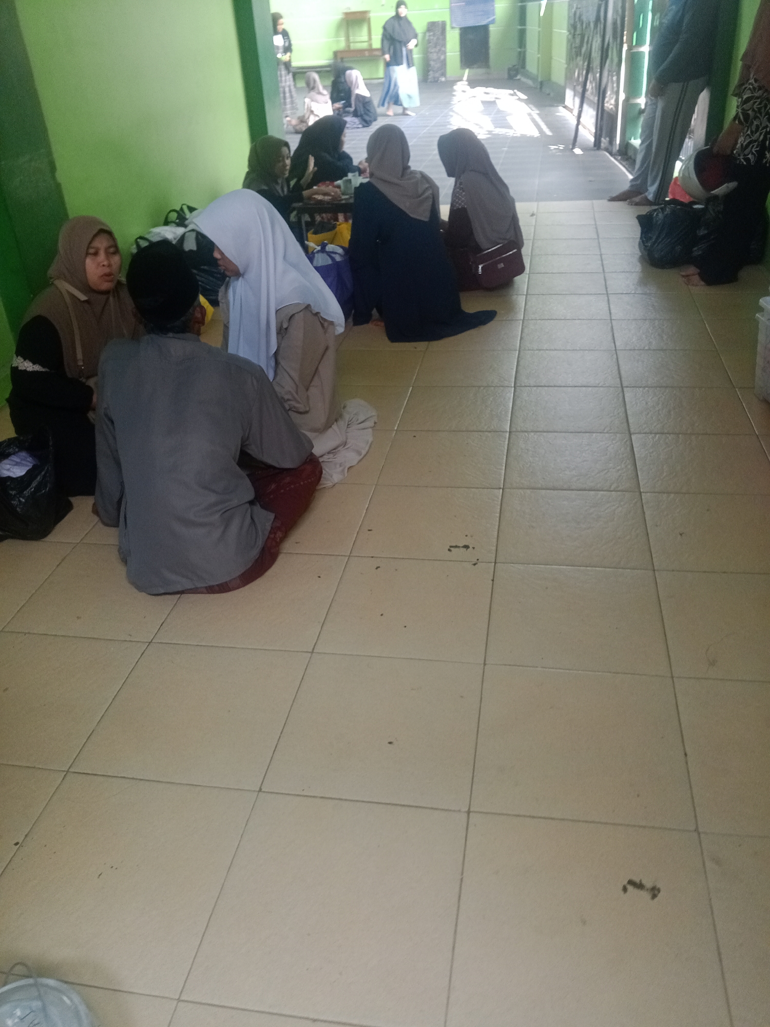 In Banyudono, Indonesia, a group of people are seated on a tiled floor in front of a light green wall. The scene appears to be indoors, possibly in a community hall or waiting area. Several individuals are wearing headscarves, suggesting a cultural context.
In the foreground, a person in a grey long-sleeved shirt and dark red pants is sitting on the floor, facing away from the camera. To their left, two women are seated, one in a brown headscarf and the other in a white headscarf. Their interaction is unclear, but they are positioned close to each other.
Further back, a woman in a dark headscarf is seated. To her right, another woman in a grey headscarf is also seated. The background shows more people standing and sitting, though their details are less clear due to distance and resolution. The environment includes a green wall, a tiled floor, and some bags on the floor. The lighting suggests it is daytime, with natural light possibly entering from a side opening.
