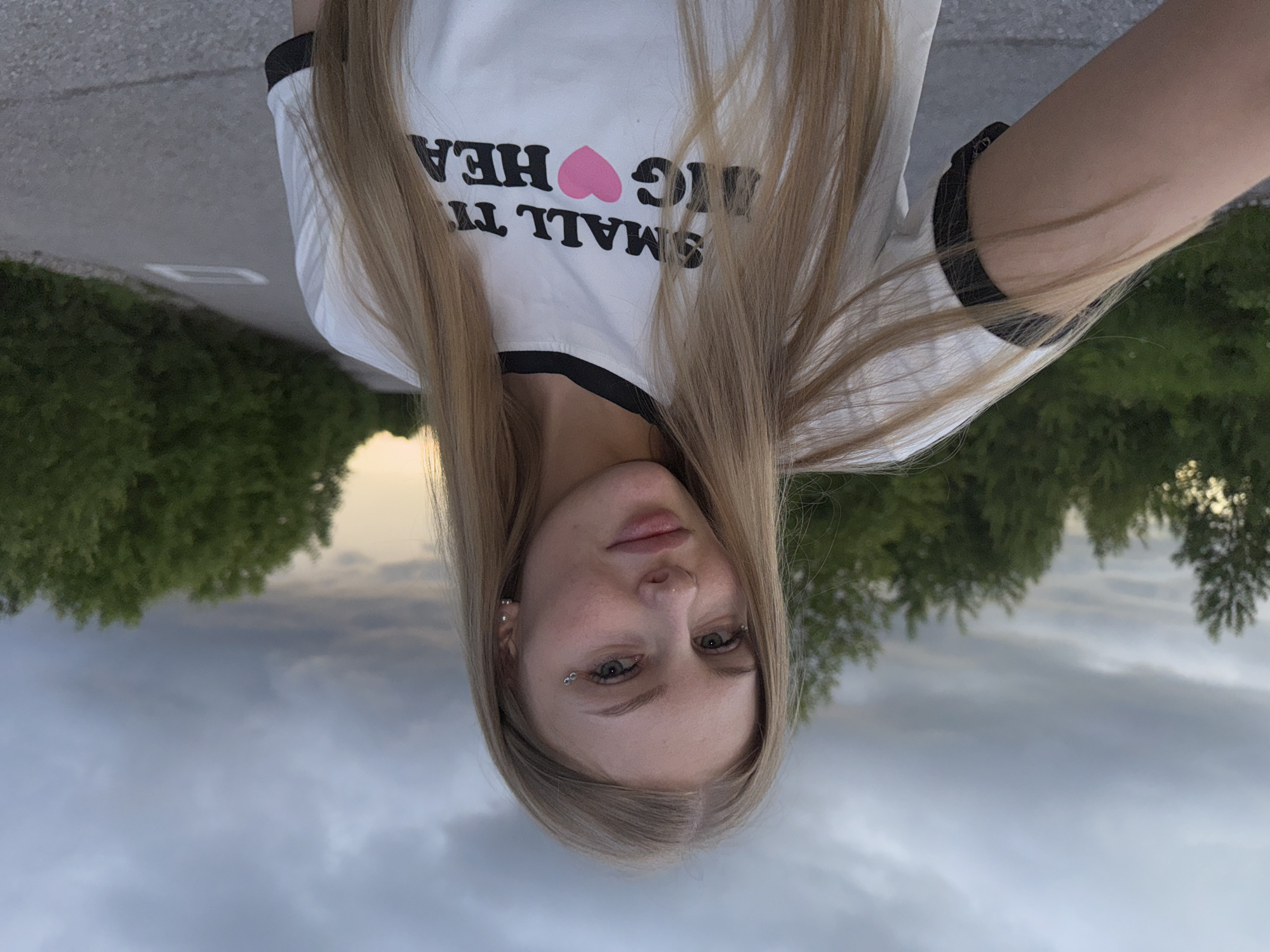 A young woman with long blonde hair and light skin poses for a selfie outdoors on a paved road. She is wearing a white t-shirt with black trim around the collar and sleeves, featuring the text "SMALL THING BIG HEART" with a pink heart. She has makeup on, including eyeliner and what appears to be glitter or gems on her face near her eye. The background consists of a cloudy sky and trees lining the road. The location is not specified as the city of Perm, Russia.