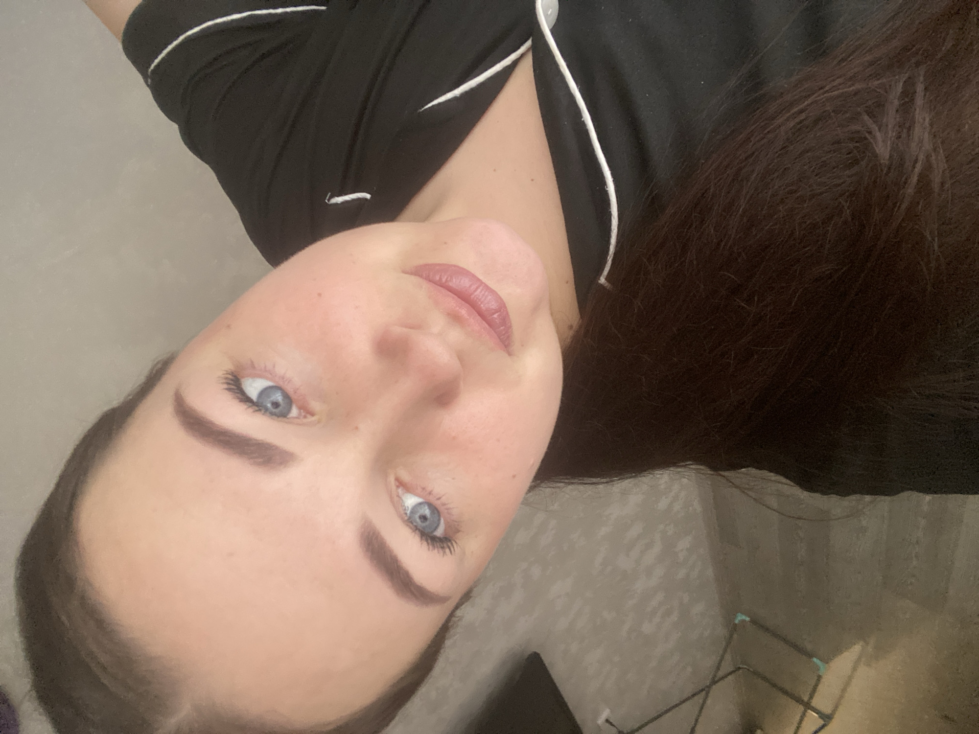 A woman with blue eyes and brown hair is looking up at the camera. She is wearing a black top with white piping. The background is blurry and shows a patterned wall and a clothes drying rack. There is no evidence to suggest the location is Sochi, Russia.