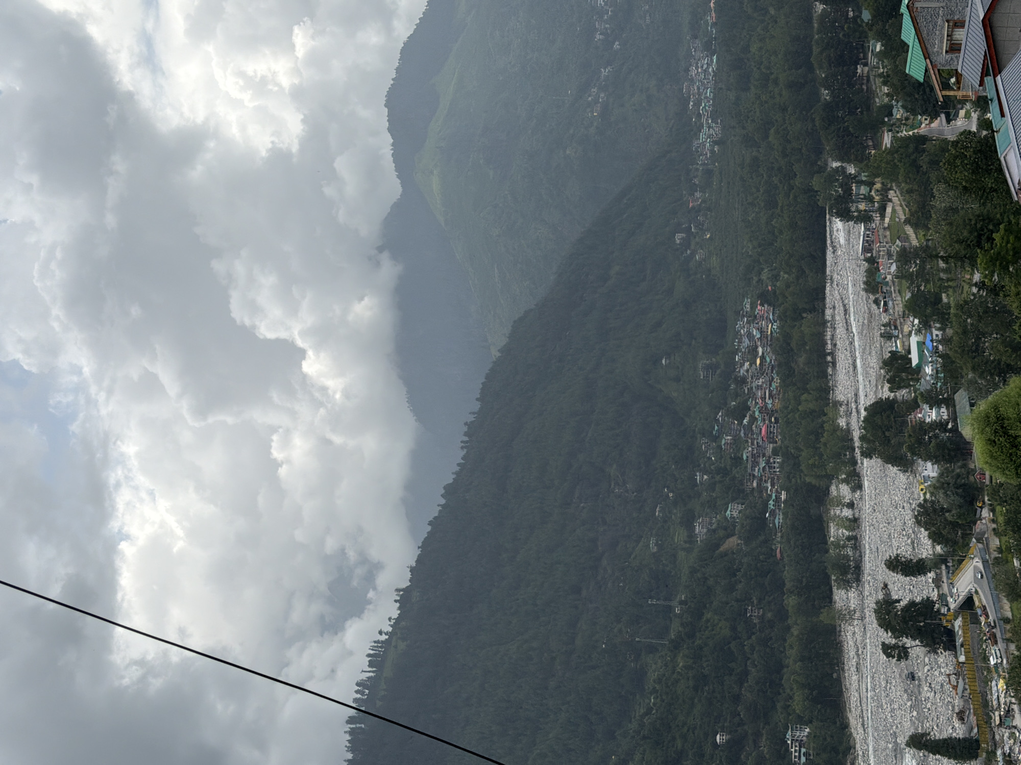 A wide shot captures a cloudy, overcast day in the mountainous region of Vashisht, India. The image is dominated by lush green mountainsides covered in dense trees. A wide, rocky riverbed runs through the valley, with a small town nestled between the mountains and the river. The town features a collection of buildings with colorful roofs, indicating human habitation. The sky is filled with heavy, gray clouds, obscuring the tops of some of the higher mountains. A black cable stretches diagonally across the upper left portion of the frame, suggesting the presence of some form of infrastructure like a cable car or power line. The overall impression is one of a serene, natural landscape under a dramatic, cloudy sky.