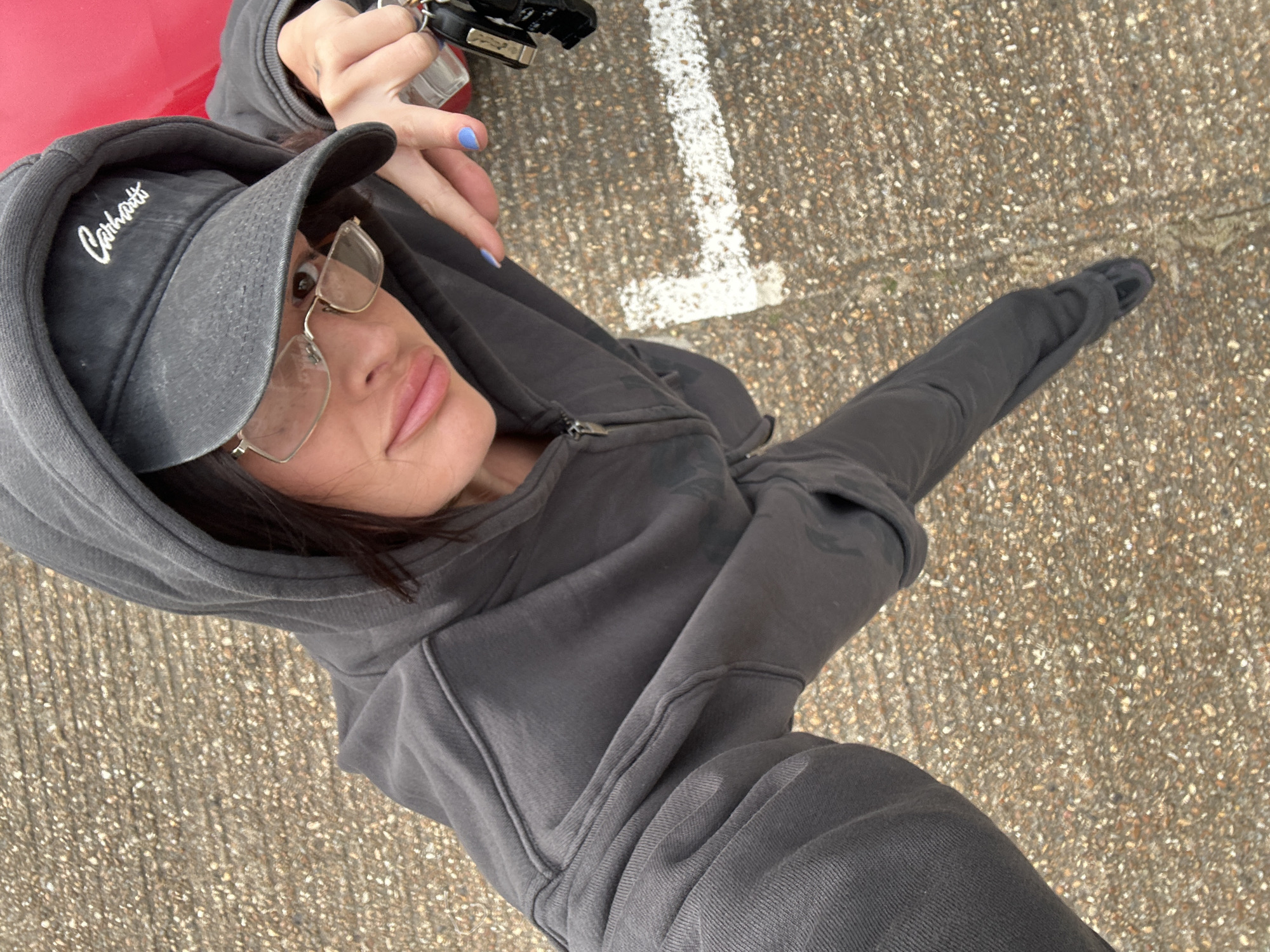 The image shows a person wearing a gray Carhartt hat, glasses, and a gray hoodie and leggings, possibly standing on a concrete surface with white markings. The person is holding car keys and the image appears to be taken from a high angle, capturing the person's face and body along with a section of the ground. The location cannot be determined from the image as it lacks identifiable landmarks or signs pointing to Tunbridge Wells, United Kingdom.