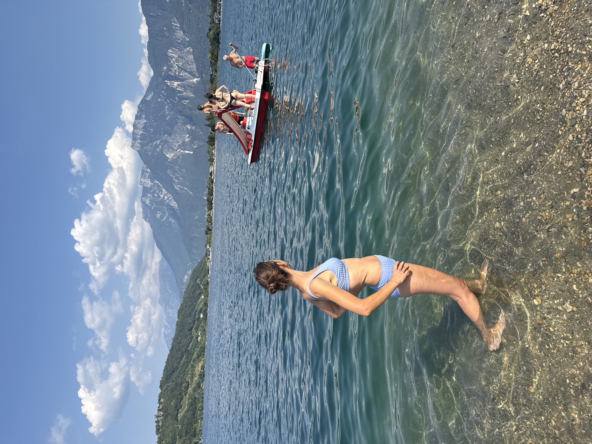 In the city of Calceranica al Lago, Italy, a woman stands waist-deep in the clear, shallow water of a lake. She is wearing a blue and white striped bikini. Her hair is pulled back in a bun. She is looking out at the lake, where a red and white pedal boat is visible in the distance. The pedal boat contains five people: three women and two men. Two of the women are standing, one is sitting, and one man is standing while another man is seated. They are on a sunny day with a clear blue sky and mountains in the background. The sun's rays are visible on the surface of the water. The lake bed is covered with pebbles.