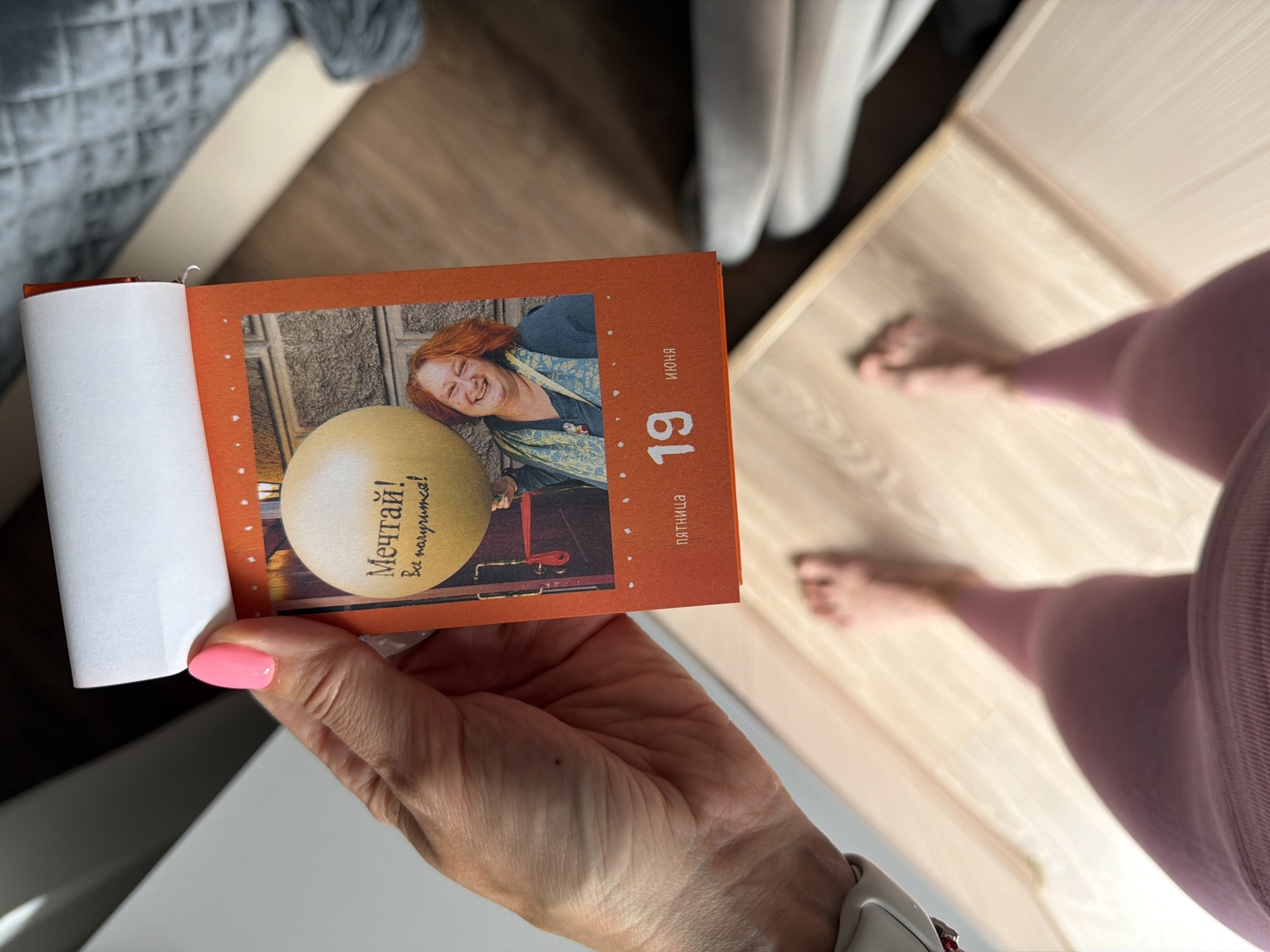 A person's hand with bright pink nail polish holds up a calendar page. The page features a photograph of a red-haired woman with a large yellow balloon and text that reads "Мечтай! Всё получится!" (Dream! Everything will work out!). Below the photograph, the date "19 ИЮНЯ" (June 19) is displayed, along with the word "ПЯТНИЦА" (Friday). The background of the image shows a blurred wooden floor and a portion of what appears to be furniture. The image is taken from a high-angle perspective, looking down at the calendar page and the person's legs and feet, who are barefoot and wearing pink leggings. The scene does not provide any specific information or context that would indicate it is located in Birobidzhan, Russia.