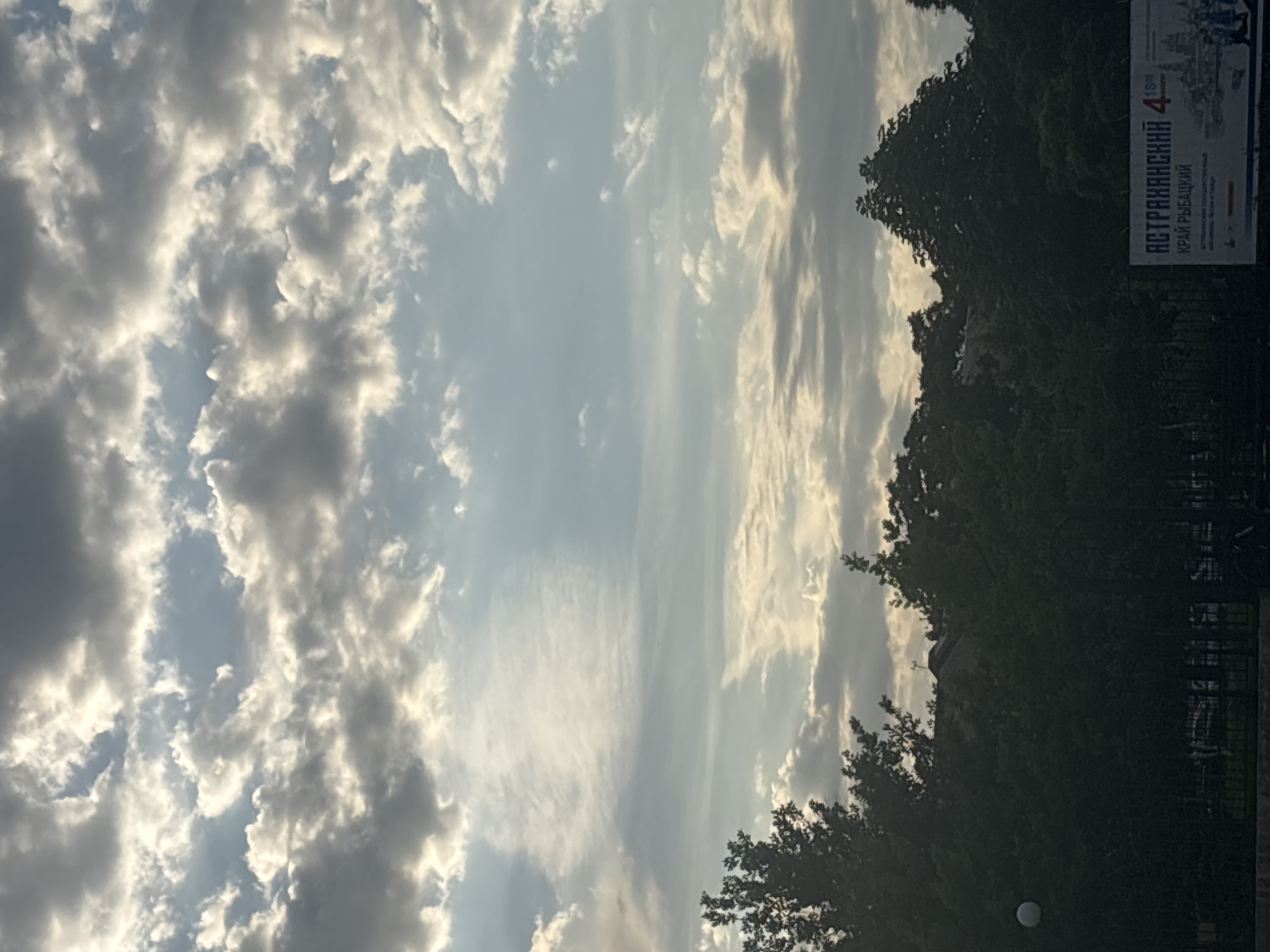 The image captures a view of the sky above a tree line in Astrakhan, Russia. The sky is filled with a mix of dark and light clouds, with sunlight breaking through in some areas, creating a dramatic effect. Below the sky, the dense foliage of trees forms a dark silhouette against the brighter sky. A billboard is partially visible in the lower right corner, featuring Russian text that appears to be related to the Astrakhan region. The text includes "АСТРАХАНСКИЙ" (Astrakhansky) and "КРАЙ РЫБАЦКИЙ" (Fisherman's Land), suggesting the billboard promotes tourism or the region's fishing industry. The overall scene conveys a natural setting with hints of urban infrastructure in the background.
