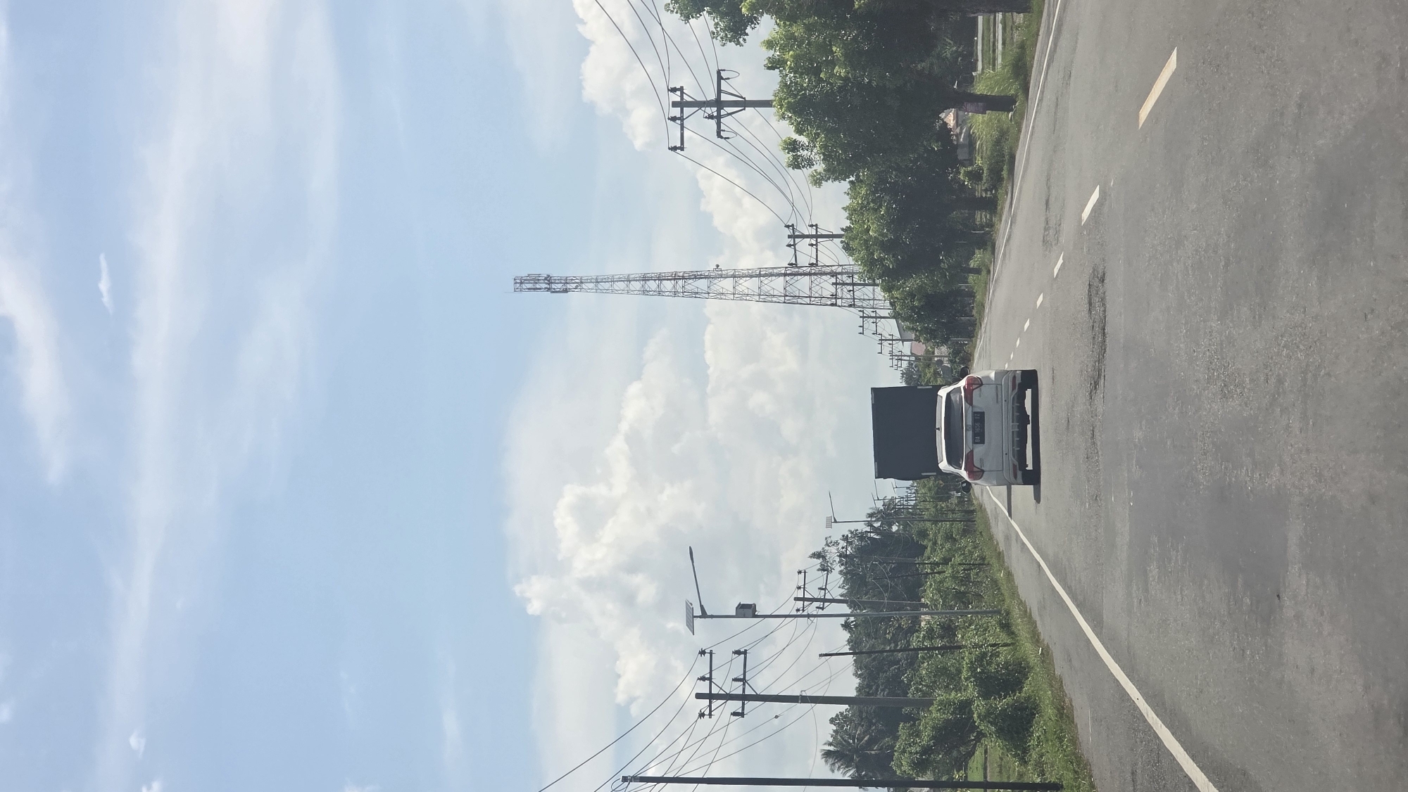 A street in Kampungjumpa, Indonesia. A white car travels on the right side of the road. Power lines run along the side of the road. In the background, a radio tower stands near cumulus clouds.