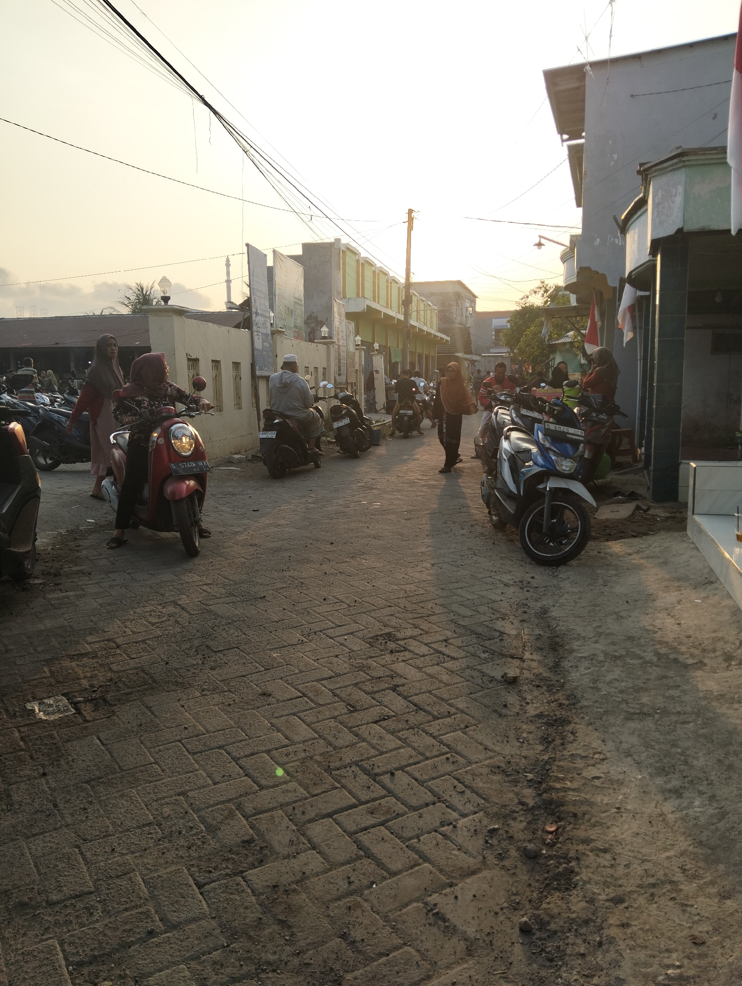 The image depicts a street scene in Banjarwati, Indonesia, during what appears to be late afternoon or early evening, indicated by the warm, low-angled sunlight and soft shadows. The street is paved with interlocking brick pavers. On the left side of the street, there are several buildings, some with green accents and others that appear to be shops or residences. A minaret of a mosque is visible in the distance.

Several people, predominantly wearing headscarves and some in traditional Islamic attire, are present. Many are either standing or seated on motorcycles. A red scooter is in the foreground on the left, with a woman wearing a red headscarf seated on it, and another woman standing beside her. Further down the street, a man in a white skullcap is riding a dark-colored scooter. On the right side of the street, there are more buildings, including one with a distinctive curved facade, and a few people are visible near the entrance.

Various motorcycles, including scooters and what appear to be more standard motorcycle models, are parked or being ridden along the street. Some are in motion, indicated by blurred tires and the presence of riders. The overall atmosphere suggests a community gathering or a busy period in the local marketplace or residential area. The sky is a pale yellow and white, with some scattered clouds. Overhead, a network of electrical wires is strung across the scene.