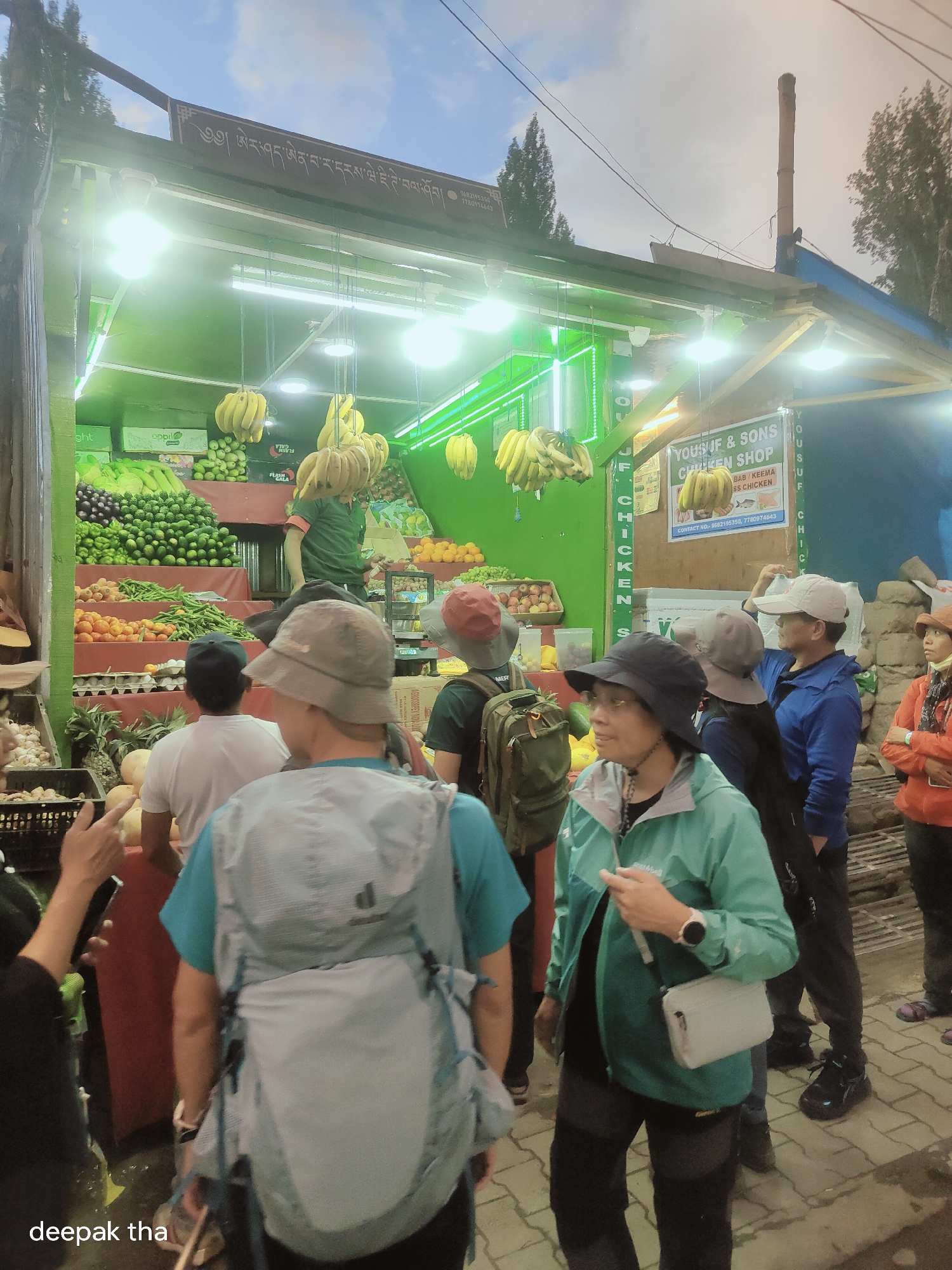 In the city of Leh, India, a brightly lit fruit and vegetable stall is the central focus. The stall is well-stocked with various produce, including bananas, green apples, oranges, and a large assortment of vegetables such as cucumbers and what appear to be green peppers. Several people are gathered around the stall, some browsing the selection and others standing in line. The scene is bustling, with an atmosphere of everyday commerce. The stall itself has a rustic appearance, with a wooden structure and neon green lighting illuminating the vibrant colors of the produce. The sky above is cloudy, suggesting either dawn or dusk. The overall impression is that of a lively marketplace scene in Leh.