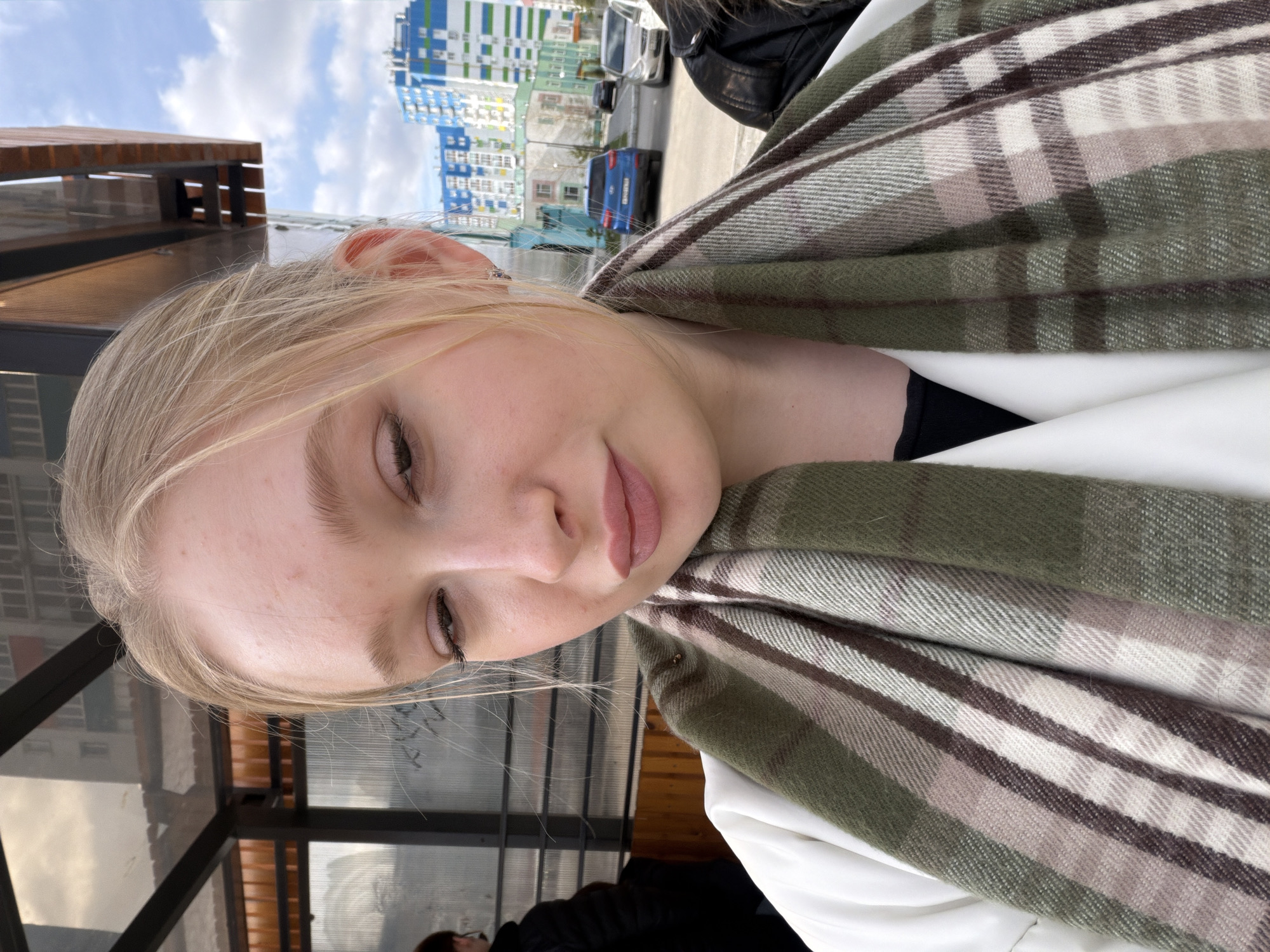 A close-up shot depicts a fair-skinned person with blonde hair wrapped in a large, striped shawl with shades of green, brown, and beige. The person is looking down, with their eyes partially closed. A white garment is visible beneath the shawl.
In the background, buildings with multicolored facades are seen on a street with parked cars. A bus stop structure with a wooden roof and metal frame is also visible, suggesting an urban environment. Given the provided location of Засечное, Russia, the scene likely captures a moment of daily life within this city.