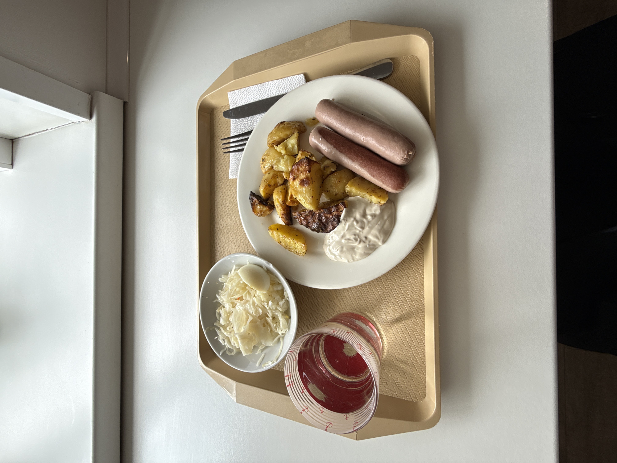 The image shows a meal tray, likely in a hospital or institutional setting. The tray contains a white plate with two sausages, roasted potatoes, and a dollop of white sauce. Beside the plate, there's a small bowl of coleslaw with a hard-boiled egg and a red plastic cup with some liquid. A fork, knife, and napkin are arranged on the upper left of the tray. The tray is placed on a white surface next to a white wall. The location is presumably in Izhevsk, Russia. No people are visible in the image.