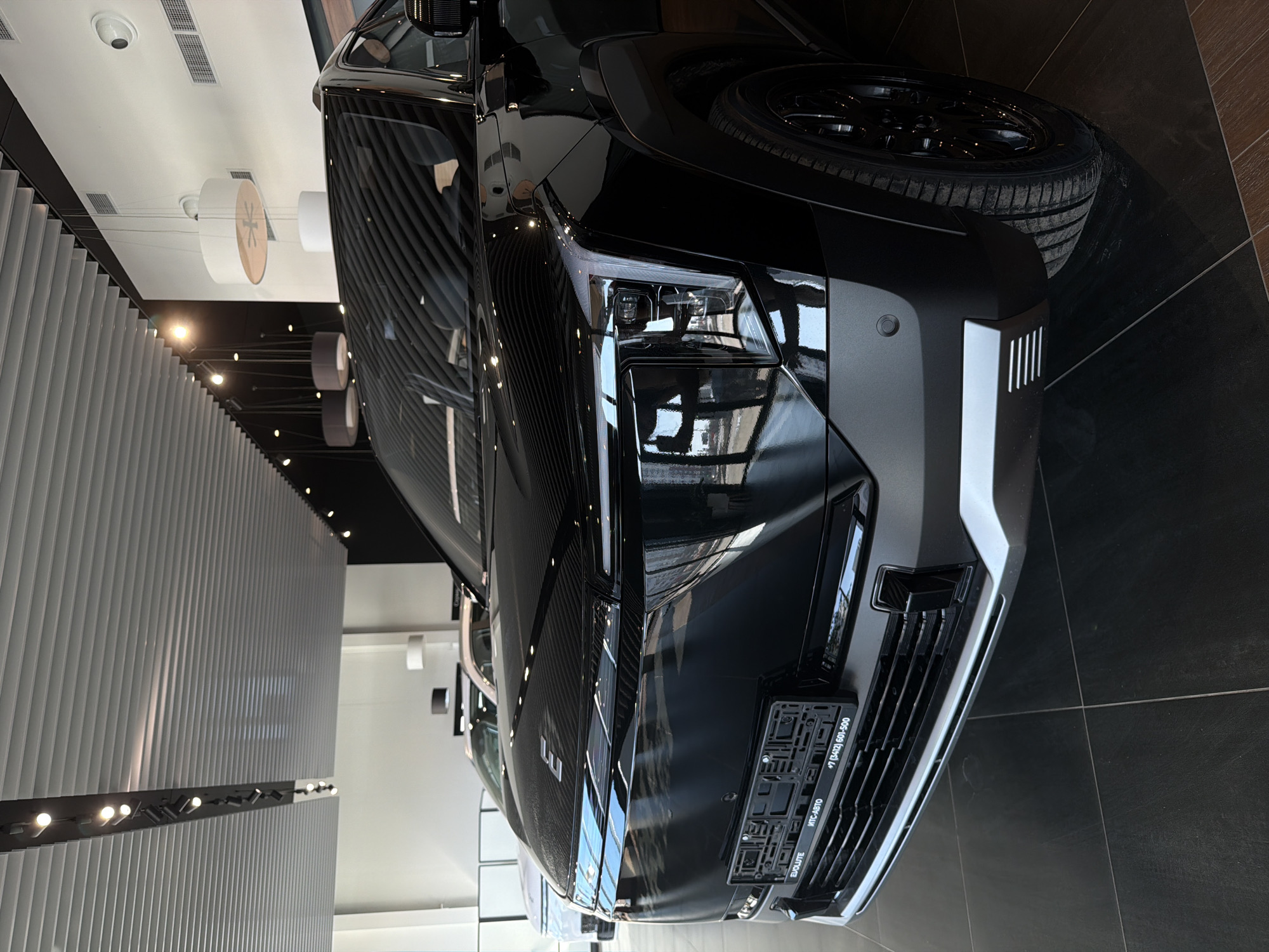 The image shows the front of a black car in what appears to be a car dealership. The car is sleek and modern, with a prominent front grille and stylish headlights. The showroom has a contemporary design with bright lighting and a clean aesthetic. A license plate holder is visible, bearing the text "EVOLUTE" and "+7 (3412) 601-500 МТС-АВТО". Based on the phone number area code, the location is Izhevsk, Russia.