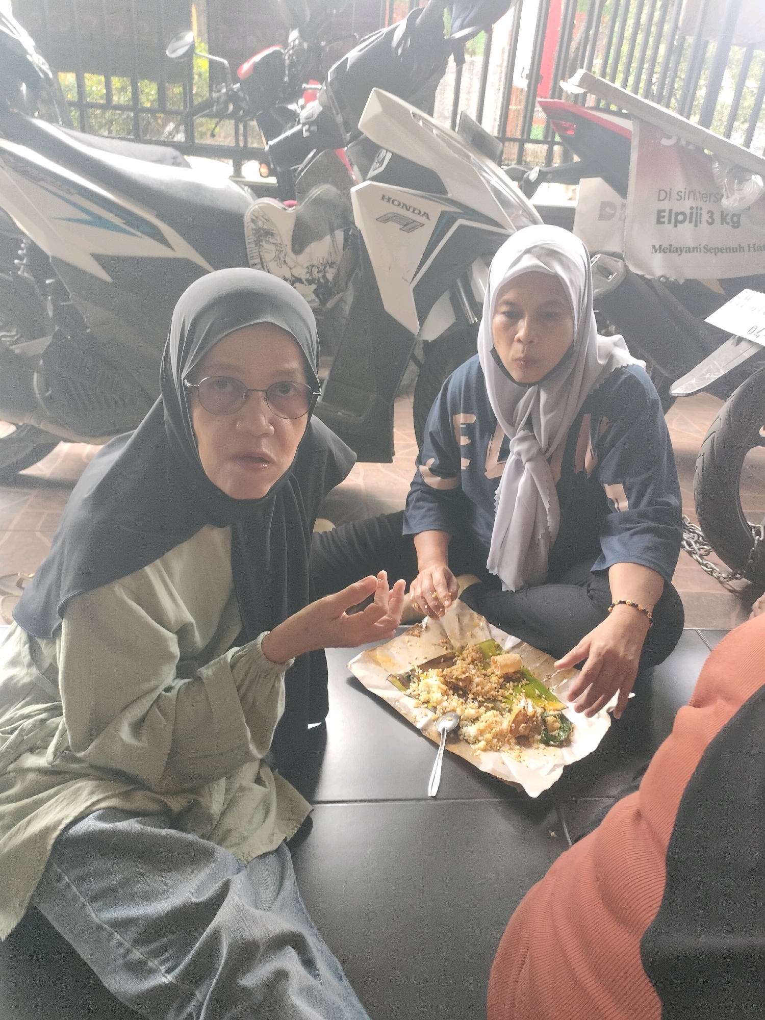 Two women are eating food from a paper wrapper on the ground. The woman on the left wears a black hijab and glasses, and is looking at the camera. The woman on the right wears a white hijab and a blue shirt, and is looking at the food. Motorcycles are parked in the background, and a metal fence is visible. The scene appears to be outdoors in an urban environment. This location is a residential street or alleyway in Pekanbaru, Indonesia, as suggested by the presence of motorcycles, modest clothing, and the likely language on the poster in the background.