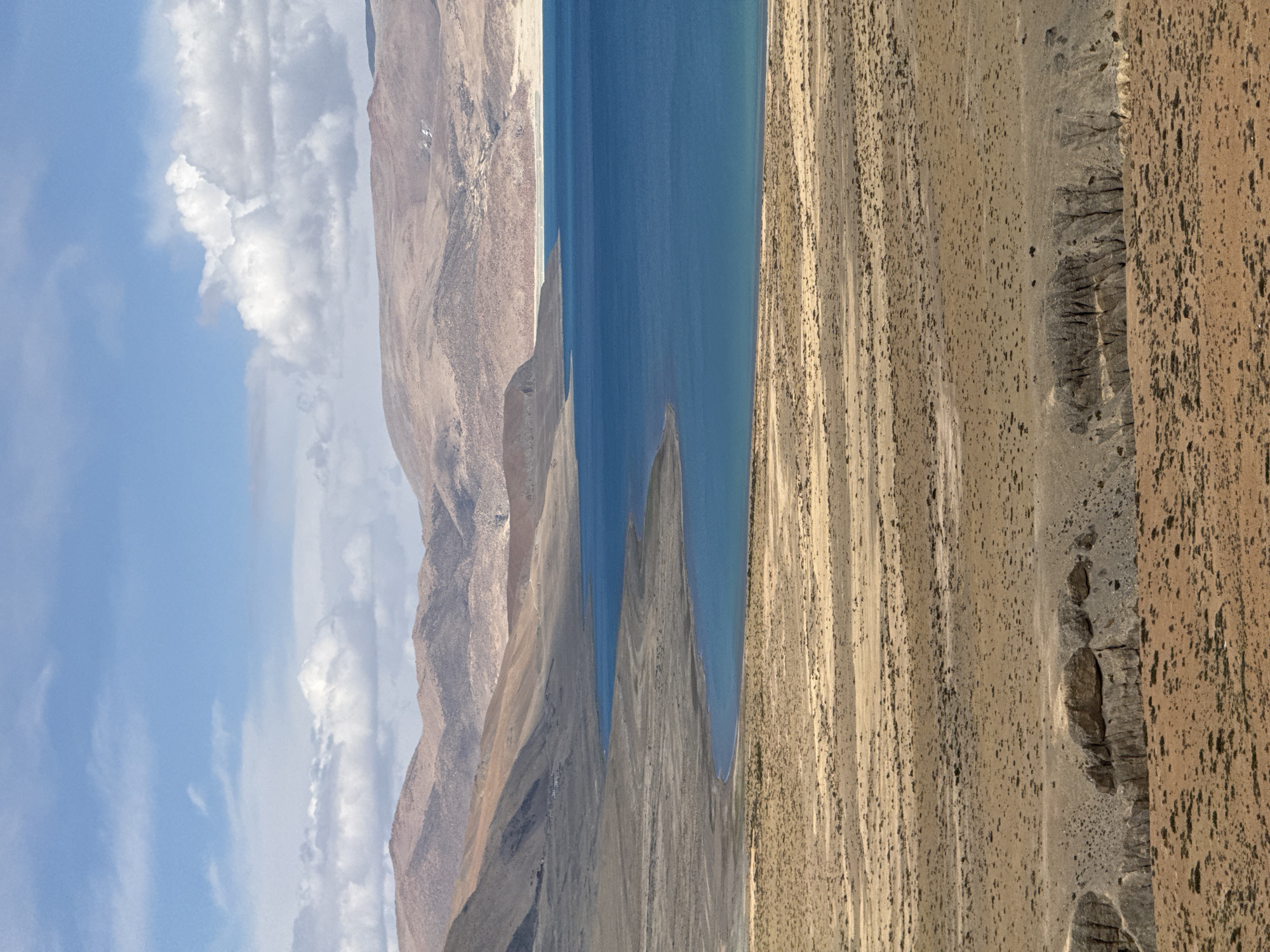 The image shows a landscape with a blue body of water vertically oriented in the middle, possibly a lake. To the left of the water is a series of brown, rocky mountain formations under a blue sky dotted with white clouds. To the right of the water is a vast, arid land with varying shades of brown and tan, suggesting desert or plateau-like terrain. The scene is likely in Gyirong County, China, given the landscape characteristics of that region. There are no people visible. The environment is dry and rugged.