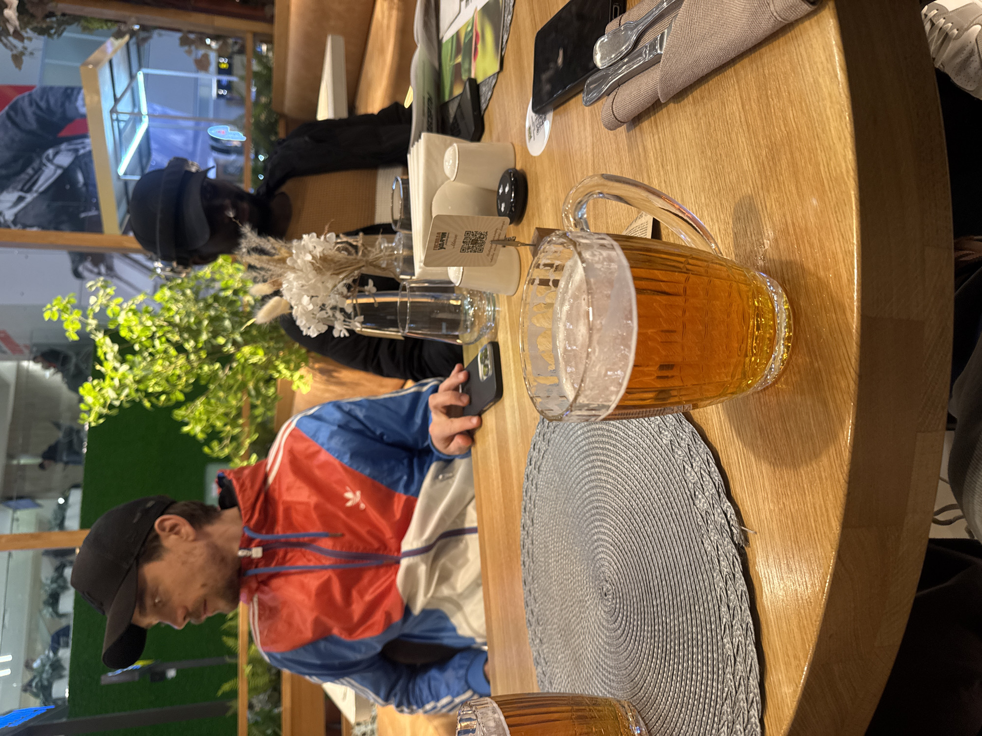 The image, correctly oriented, depicts an indoor scene at what appears to be a restaurant or bar. Two individuals are seated at a light wooden table. In the foreground, a man wearing a blue, red, and white Adidas jacket with a Trefoil logo and a black baseball cap is looking down, holding a smartphone. Adjacent to him on the table is a large mug filled with an amber beverage, likely beer, topped with foam, resting on a round, grey woven placemat. Another beer mug is partially visible at the bottom edge of the frame.
The table is set with various items including a napkin dispenser, a small vase containing dried white flowers and foliage, empty drinking glasses, and salt and pepper shakers. A stand with a QR code is prominent on the table, displaying the text "ULTRA VARV menu". Silverware, wrapped in a grey napkin, is also visible. In the background, another person, largely obscured, is seated, wearing a black top and a black baseball cap. Green plants and reflections in glass panels suggest a decorated interior environment. The lighting indicates an indoor setting, possibly evening, with artificial illumination. The overall atmosphere is casual.