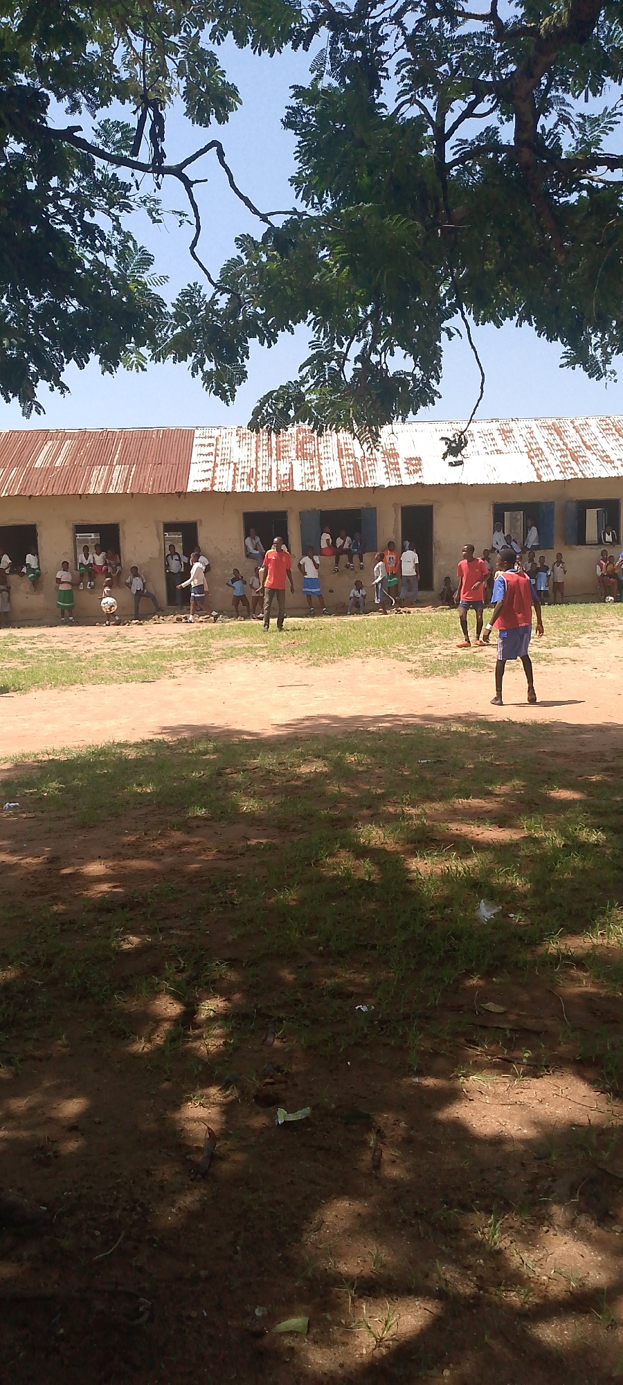The image shows a group of children and adults outside a simple, light-colored building with a corrugated metal roof. The building appears to be a school or community center, with several open doorways and windows. Many children are seated on the windowsill and inside the building, while others are on the ground outside. Some of the children and at least two adults are playing soccer in the dirt yard in front of the building. The weather appears to be sunny and clear, with shadows cast by trees in the foreground. The overall scene suggests a casual, outdoor activity taking place during the daytime. There is no visible text in the image.
