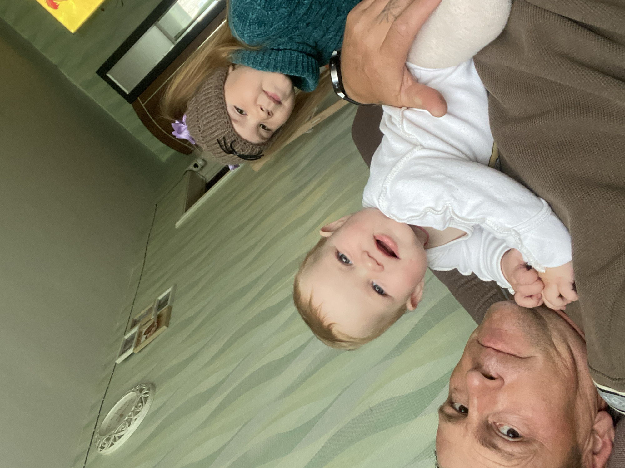 This image depicts three individuals in a domestic setting, located in the city of Фоки, Russia. An adult male with short brown hair and light skin is positioned in the foreground, lying on his back and looking directly towards the camera. His right arm is visible, featuring a dark wristwatch. An infant with light blonde hair and blue eyes, wearing a white long-sleeved top and light leggings, is lying prone on the adult male's chest, facing the camera with an open-mouthed expression. A young girl with long light brown hair, wearing a brown knitted hat with black ear-like protrusions and a teal turtleneck sweater, is positioned behind the infant, also looking towards the camera with a slight smile. Her hair is tied with a purple accessory. The individuals are situated on a textured brown fabric, likely a couch or bed. The background consists of a wall covered with light green wallpaper featuring a wavy, striped pattern. On the left section of the wall, an oval-shaped ornate white decorative piece and a multi-frame picture collage are visible, with a dark cable running horizontally above them. On the right section of the background, a bright yellow square art piece with a red element is visible above a dark-framed doorway.