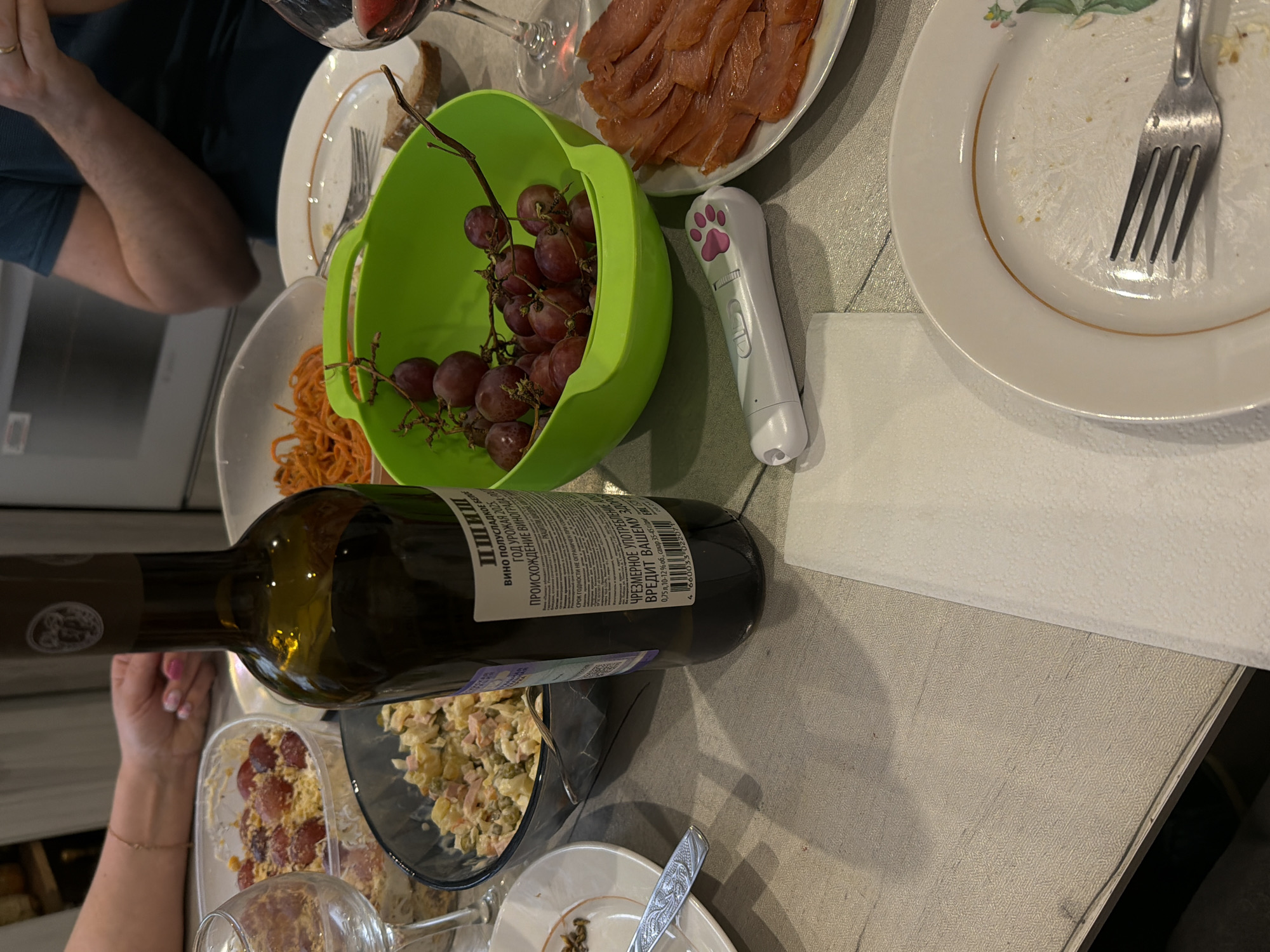 The image captures an indoor dining scene, likely a social gathering or meal, set in Reshetnikovo, Russia. The perspective is an overhead view of a table laden with food and drinks.
Visible subjects include portions of people, indicated by arms and hands, one wearing a ring and another with painted nails. The table is set with several dishes: a vibrant green bowl filled with red grapes, a white plate piled with sliced cured fish (possibly salmon), and a bowl containing what appears to be Olivier salad. Another bowl holds an orange-colored dish, resembling noodles or shredded carrots. Empty or partially eaten white plates, one with a fork, suggest the meal is in progress or recently finished. A dark glass wine bottle lies across the table, obscuring some dishes and a wine glass. A distinctive white gadget with a pink paw print design is also on the table.
The wine bottle features a label with Cyrillic text. Recognizable phrases include "ВИНО ПОЛУСЛАДКОЕ" (Semi-sweet wine), "ГОД УРОЖАЯ 2025" (Harvest year 2025), "РКАЦИТЕЛИ" (Rkatsiteli, a grape variety), and a warning about "ЧРЕЗМЕРНОЕ УПОТРЕБЛЕНИЕ ... ВРЕДИТ ВАШЕМУ ЗДОРОВЬЮ" (Excessive consumption... is harmful to your health). The label also states 0.75L volume and 10-12% alcohol by volume. The overall ambiance suggests a casual yet festive occasion.