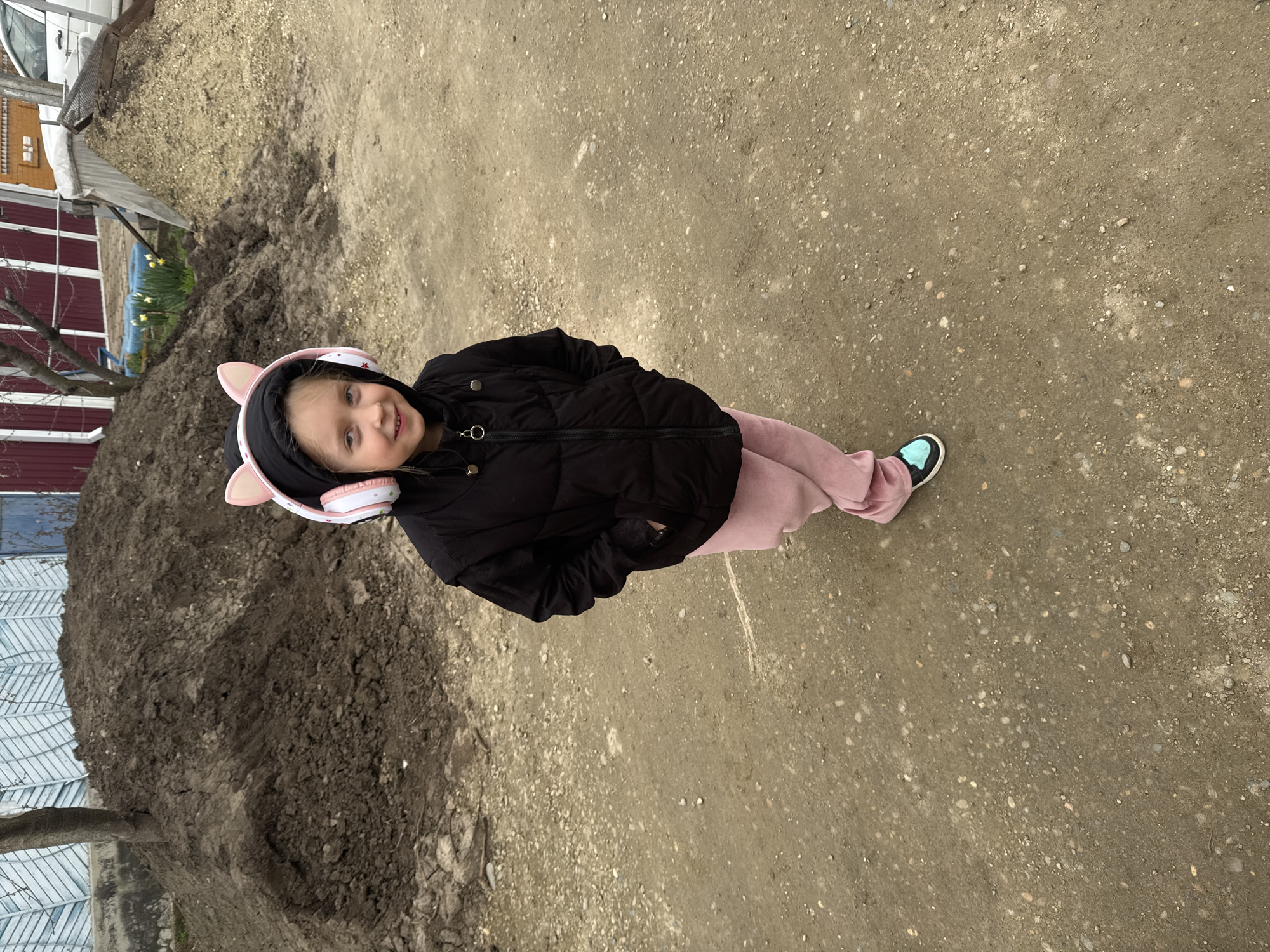 A young girl, approximately 5-8 years old, stands centrally on an unpaved dirt and gravel path in Kurganinsk, Russia. She wears a black hooded puffy jacket, pink sweatpants, and light blue and black sneakers. Pink and white cat-ear headphones are positioned over her head. Her hands are placed inside her jacket pockets, and she displays a slight smile. To her left, in the background, is a large mound of dark brown earth, partially obscuring a light blue wooden fence and a slender, bare-branched tree. To her right, a pile of lighter, sandy-colored material is visible. Behind these mounds, a red-painted fence or building wall is present, with blooming yellow daffodils. A white sedan is partially visible in the far right background.
