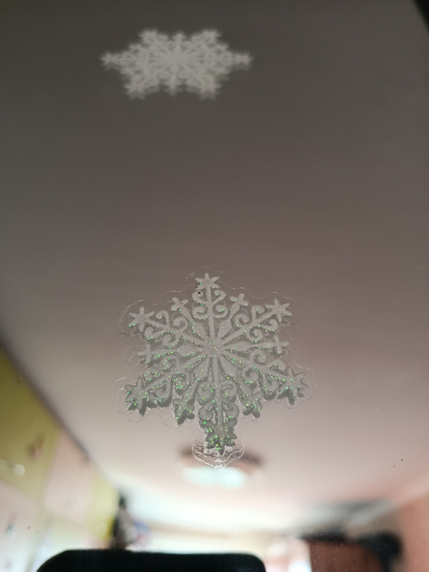 The image shows two snowflakes, one in focus and the other out of focus and reflected. The focused snowflake is on a reflective surface, possibly a mirror or window, and is adorned with green glitter. In the background, out of focus, there appears to be a room with some furniture. There are no people visible in the image. The image was taken in Biysk, Russia.