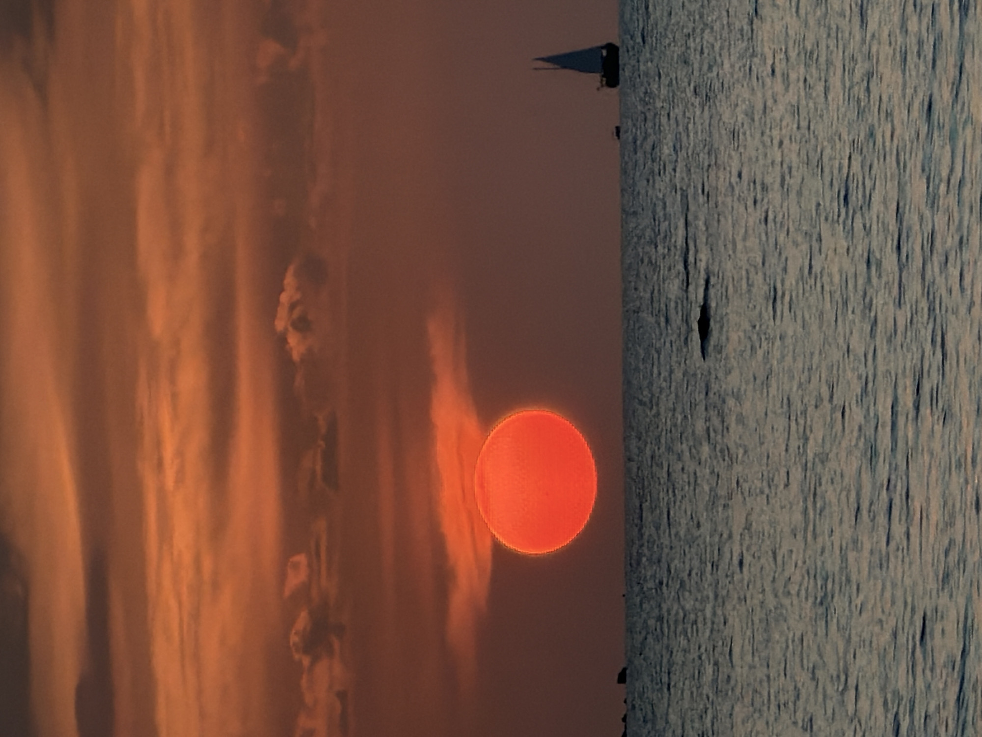 The image depicts a sunset over the sea, likely in Ko Samui District, Thailand. A large, vibrant orange sun is partially obscured by reddish-orange clouds, casting a warm glow across the sky. The ocean surface is rippled, reflecting the colors of the sunset. On the horizon, a sailboat with a single white sail is visible, along with a smaller boat. In the distance, to the left of the sun, a few dark rocks or small islands emerge from the water. The overall scene is serene and atmospheric, characteristic of a tropical sunset.
