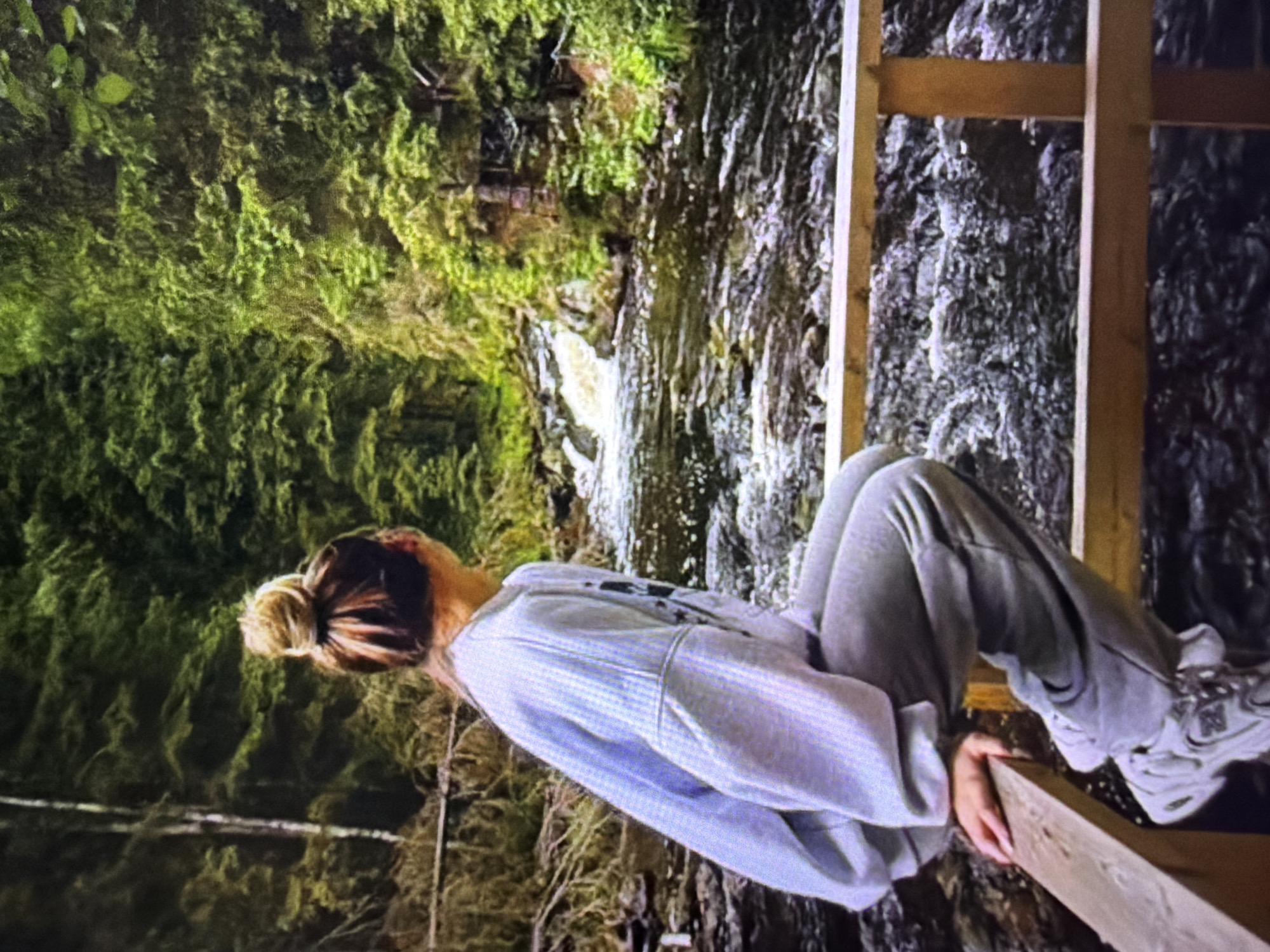 A young woman with her hair in a bun is sitting on a wooden railing, looking out at a river. She is wearing a light blue sweatshirt and grey sweatpants. The river is flowing through a lush green forest, with trees on both sides of the bank. The water is a mix of white and dark grey, indicating rapids and depth. The woman's posture suggests a moment of contemplation or observation. The scene appears to be a natural, outdoor setting, possibly a park or a hiking trail. There is no information in the image that suggests the location is in the city of Beșalma, Moldova.