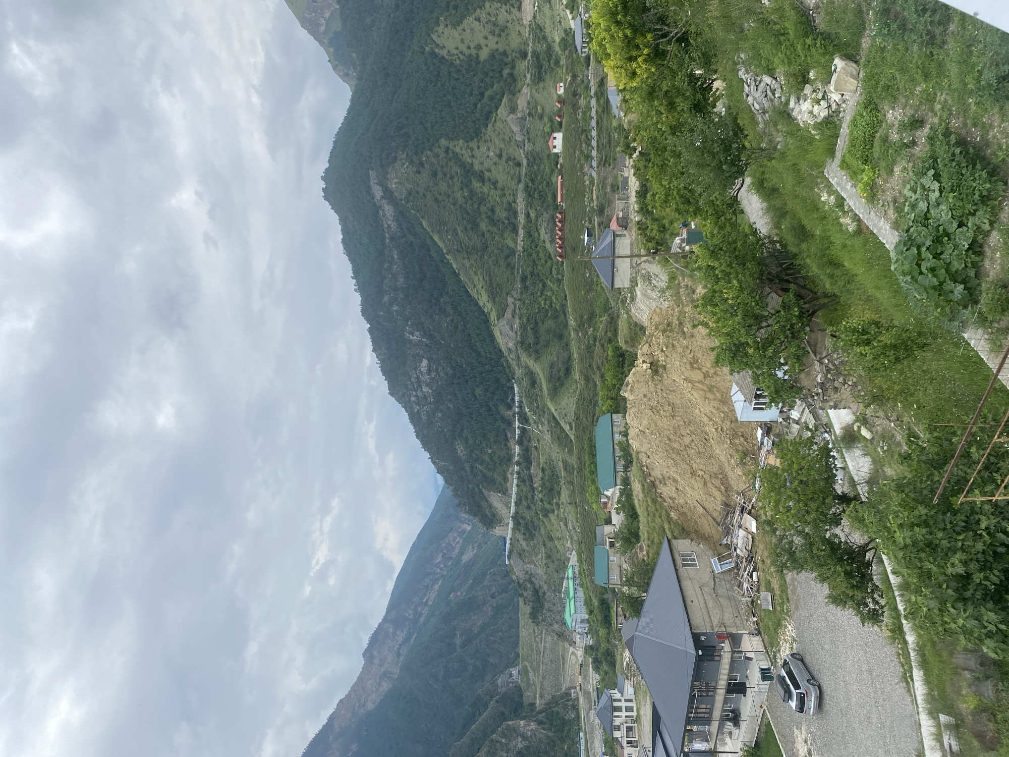 The image depicts a village nestled in a valley, framed by steep, verdant mountains. The sky is overcast with heavy, gray clouds. In the foreground, several houses are visible, some with dark pitched roofs, others with lighter colored facades. A silver car is parked in a gravel driveway next to one of the houses. A pile of construction debris, including wood and building materials, is present near a light blue house. The midground shows more houses with green roofs and some smaller structures, possibly guest houses or cabins, arranged in a row on a hillside. The background is dominated by the imposing mountains, which are densely covered with trees on their lower slopes and bare rock faces higher up. A road or path winds its way along the mountainside in the distance. The overall impression is one of a remote, mountainous settlement in a natural, rugged landscape. The image is identified as being taken in the city of Chalda, Russia.