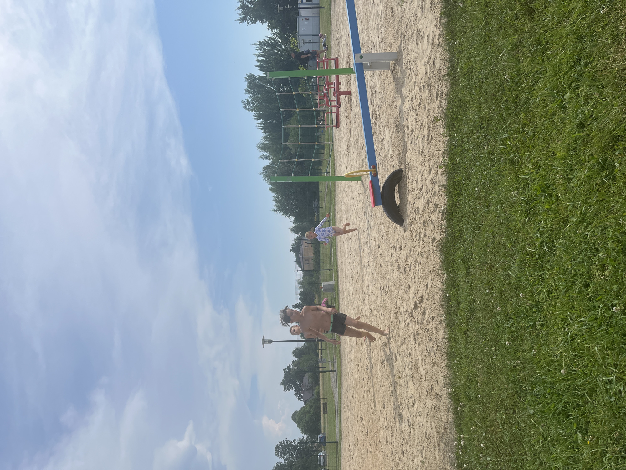 A sunny day is depicted at a sandy playground with lush green grass bordering the sand. In the foreground, a seesaw with a blue and yellow beam rests on a tire and metal supports. Behind it, a climbing net structure with green poles and netting stands tall. Several children are visible on the sand. One boy with gray hair, wearing dark shorts, runs towards the left. To his left, another boy in dark shorts is visible. Further in the distance, a young girl in a white and blue patterned swimsuit runs towards the right. A man in a dark shirt and shorts walks in the background near a building. Trees line the perimeter of the playground, and a cloudy sky with blue patches is overhead.