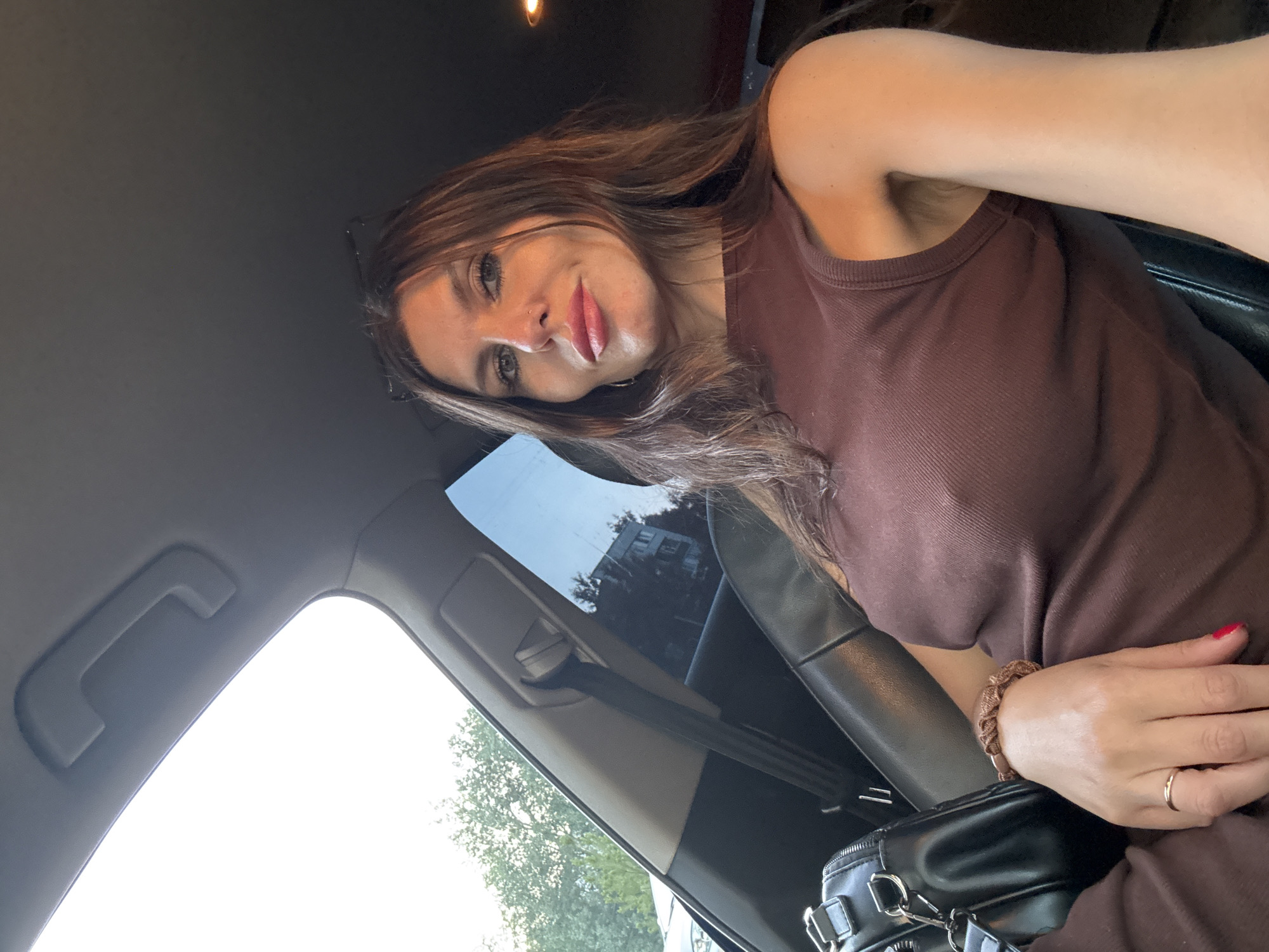 A woman is pictured inside a car, seated in the driver's seat, looking towards the camera. She has long, dark brown hair, and her makeup is visible, including lipstick and eyeliner. She is wearing a brown, sleeveless top, and a black handbag is visible to her left. A ring is on her left hand, and her fingernails are painted red. The interior of the car, including the dashboard and a seatbelt, is visible. Outside the car windows, trees and a building can be seen, suggesting an urban or suburban setting. The image was taken in Chaikovsky, Russia.