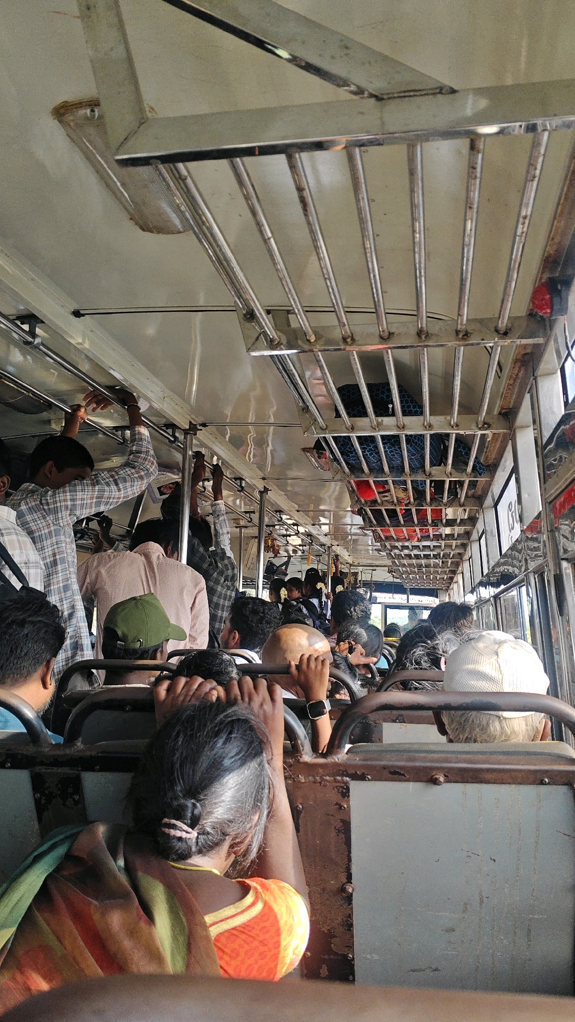 The image captures the interior of a crowded bus, possibly in Srinagar, India, with passengers standing and seated. The lighting appears to be from overhead fixtures. A metal luggage rack is visible above the heads of the passengers. The overall impression is one of congestion and a typical public transport scene in a busy Indian city.