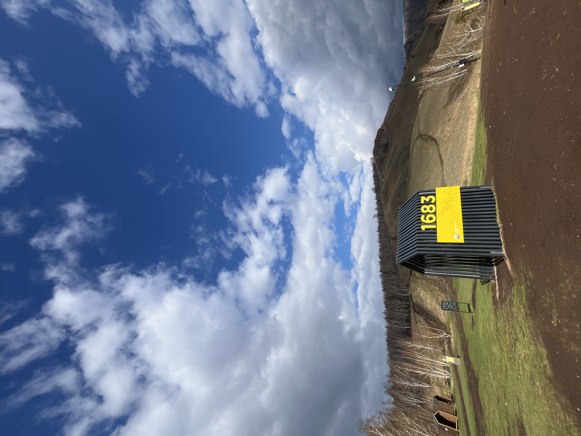 The image shows a landscape with a blue sky and clouds in the upper left corner. The bottom and right sides of the image depict a grassy area with patches of dirt. A black metal structure with a yellow sign reading "1683" is visible on the right side. The background shows a hillside with sparse trees. The location is likely in or near Krasnoyarsk, Russia, as indicated by the context.