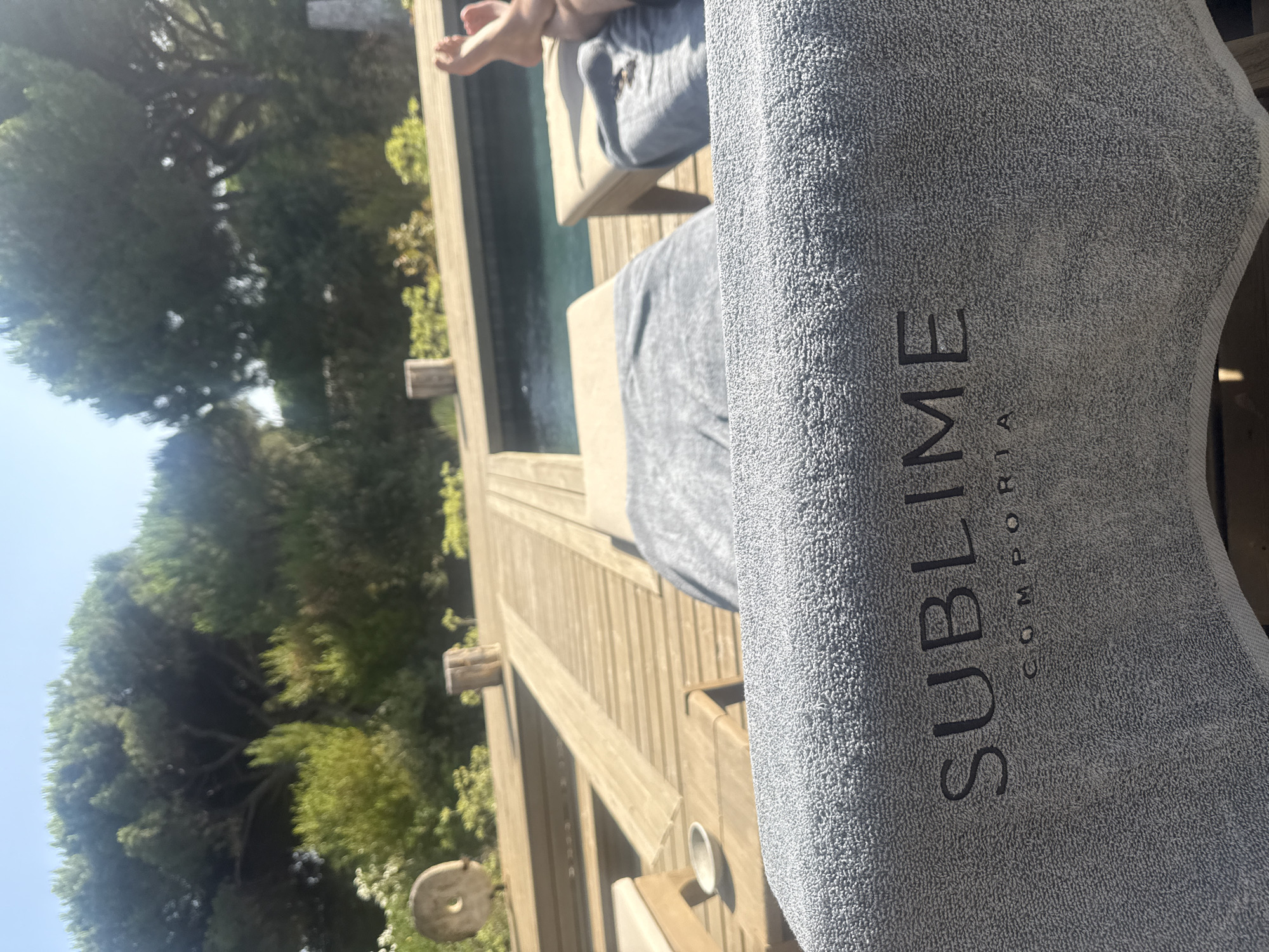 The image depicts a poolside scene at a resort in Grândola, Portugal. In the foreground, a gray terrycloth towel with the embossed words "SUBLIME" and "COMPORTA" rests on a lounge chair. Beyond the towel, a wooden deck surrounds a swimming pool. Two lounge chairs are visible on the deck, one covered with a gray towel and the other with a person's legs and feet visible, suggesting they are relaxing. Trees and foliage line the background, indicating a natural, outdoor setting. The lighting suggests it is a sunny day.