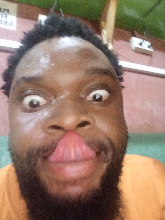 A close-up selfie captures a Black man with a full beard and dark hair. He is making a goofy face by crossing his eyes and sticking his tongue out. The overhead lighting is somewhat harsh, causing a glistening effect on his forehead. His orange shirt contrasts with the light pink and green wall behind him. An electrical outlet is visible in the upper right corner.